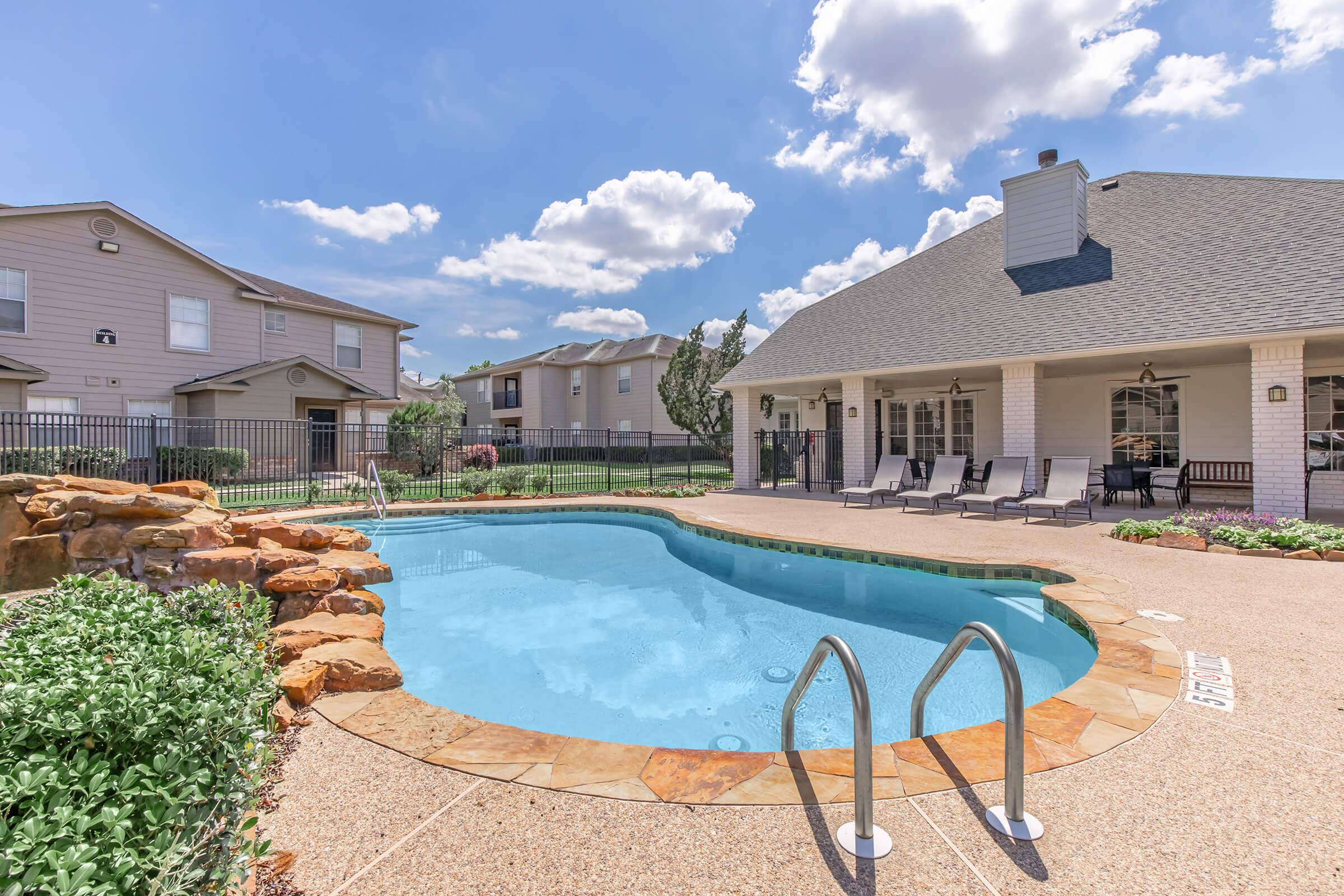 A swimming pool surrounded by lounge chairs and a landscaped area, with a backdrop of residential buildings and a blue sky dotted with clouds. The pool features a stone edge and stairs leading in, creating a welcoming outdoor space for relaxation and leisure.