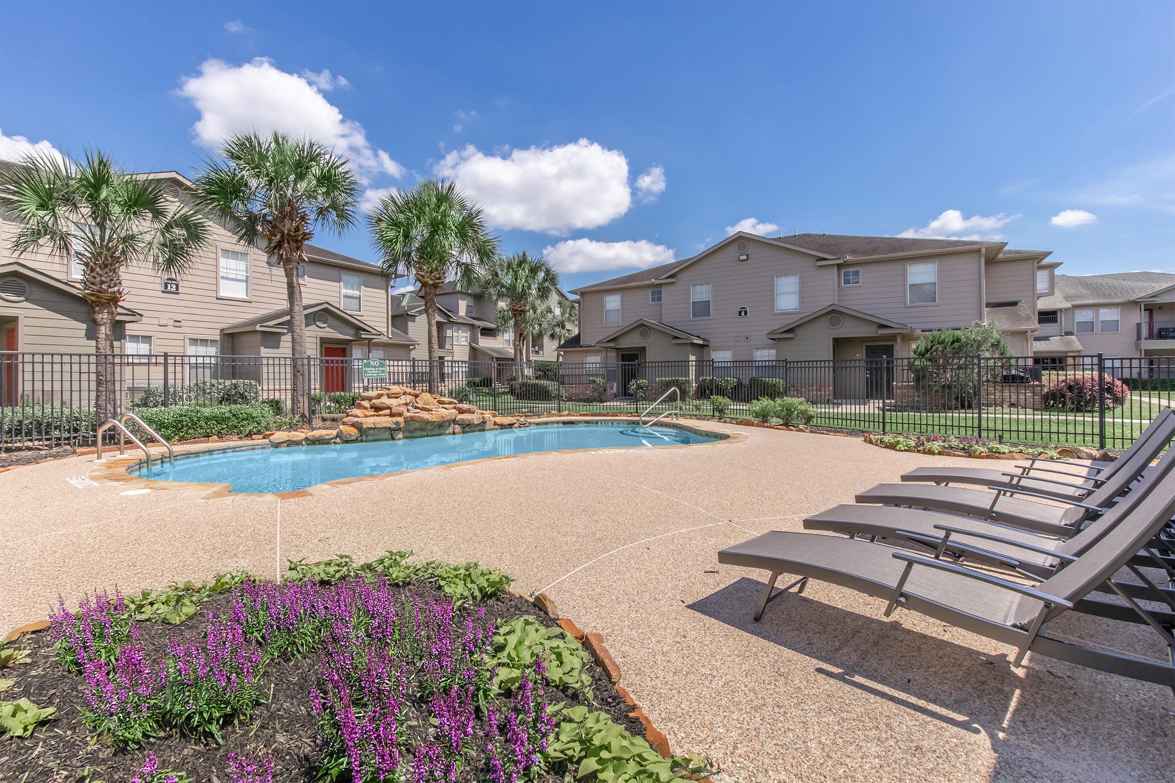 A serene swimming pool area surrounded by palm trees and vibrant flowers, with lounge chairs positioned nearby. The scene captures a sunny day, with well-maintained apartment buildings in the background, creating a relaxing outdoor environment for residents.