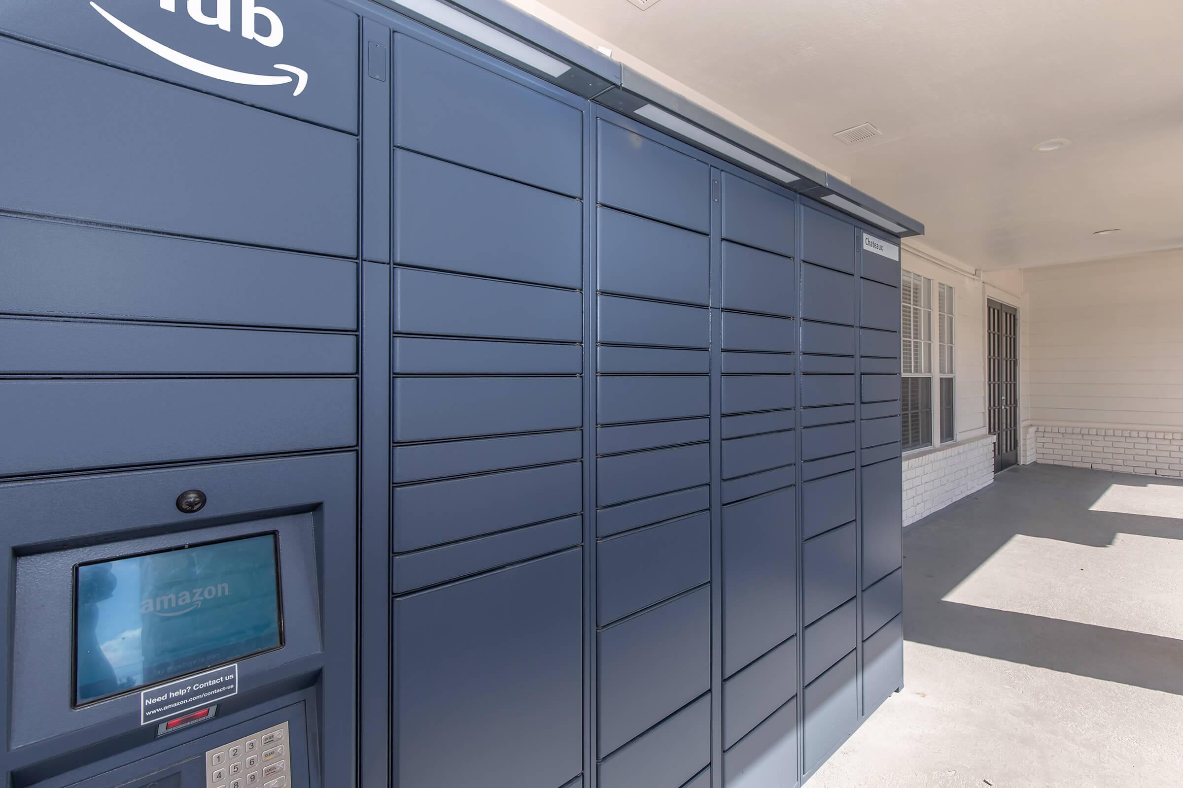 Amazon delivery locker unit installed outside a building, featuring several compartments for package pickup. There is a digital interface at the bottom displaying the Amazon logo. The setting appears to be a covered area with a concrete floor and a door leading to a nearby building.