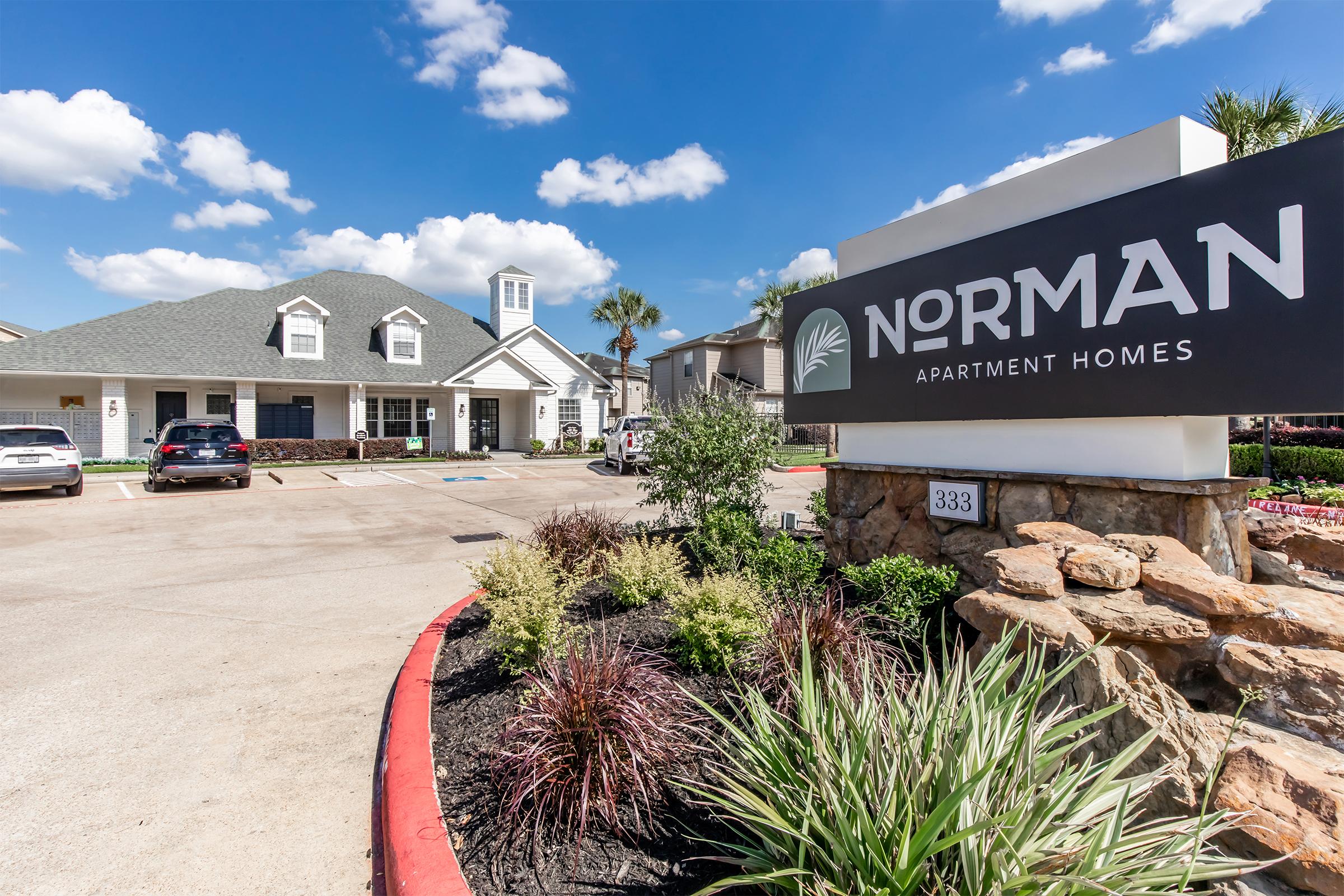 Exterior view of Norman Apartment Homes featuring a modern building with a peaked roof and a welcoming entrance. The property is landscaped with greenery and flowers. A large sign displays "NORMAN" prominently, and several parked cars are visible in the parking lot under a bright blue sky dotted with fluffy white clouds.