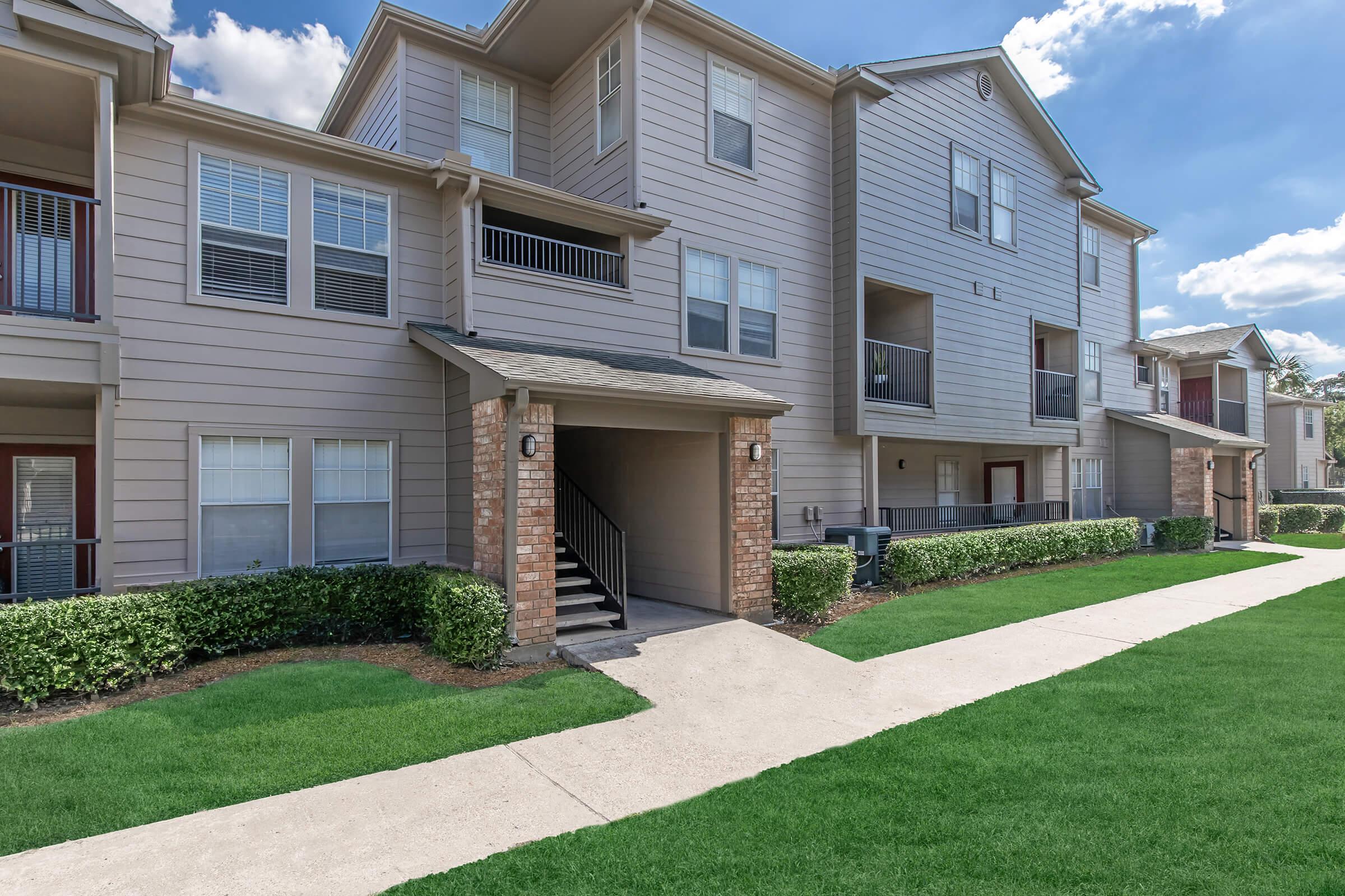 A multi-unit apartment building with beige siding and brick accents. The exterior features multiple windows, a staircase leading to upper units, and manicured green lawns. A concrete pathway runs along the front, with shrubs lining the edges. The sky is clear with fluffy clouds.
