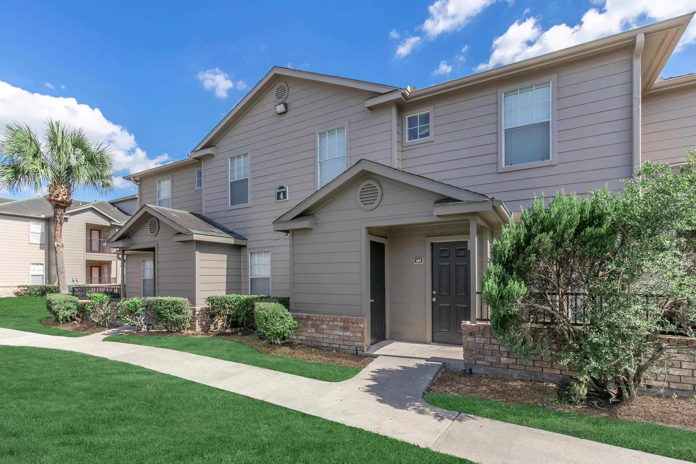 Two-story residential building with light brown siding and dark doors. Well-maintained landscaping features green lawns and shrubs. The sky is bright blue with fluffy white clouds. Pathway leads to the entrance, creating a welcoming atmosphere.