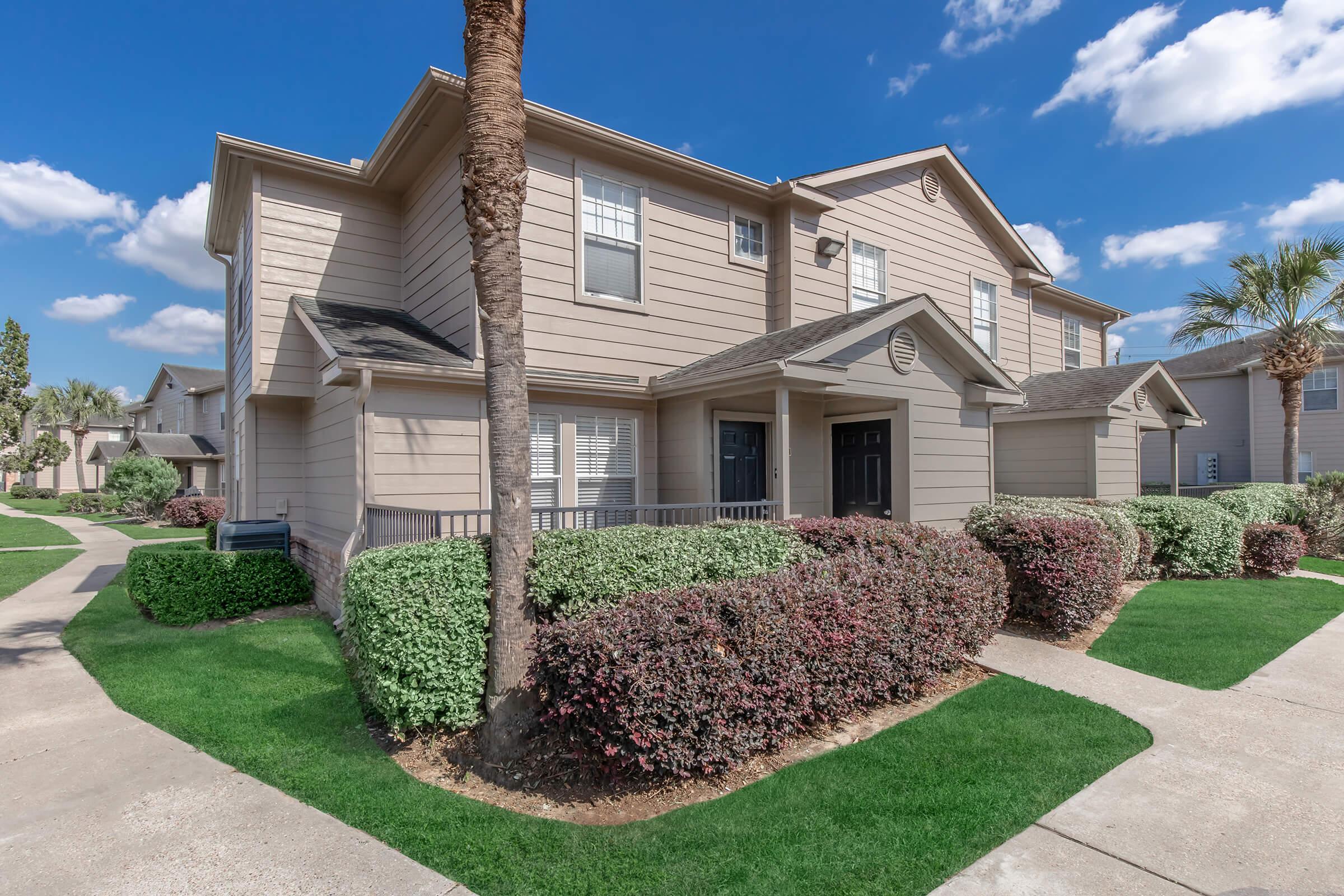 A residential complex featuring two-story beige buildings with balconies. The landscape includes well-maintained green lawns, trimmed bushes, and palm trees, under a clear blue sky with scattered clouds. Walkways connect the units, creating a welcoming outdoor atmosphere.