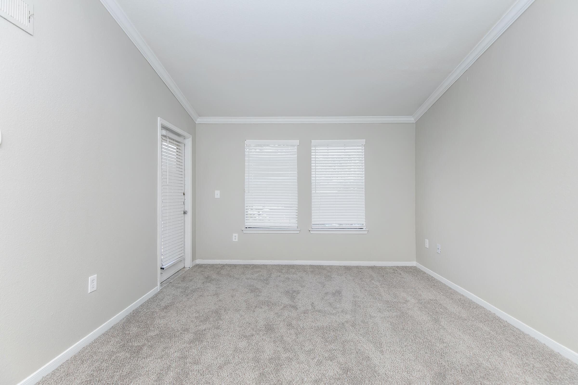 A spacious, empty room with light gray walls and soft, beige carpeting. Two windows with white blinds allow natural light to enter, while a door on the left leads outside. The ceiling features white crown molding, creating a clean and modern aesthetic.
