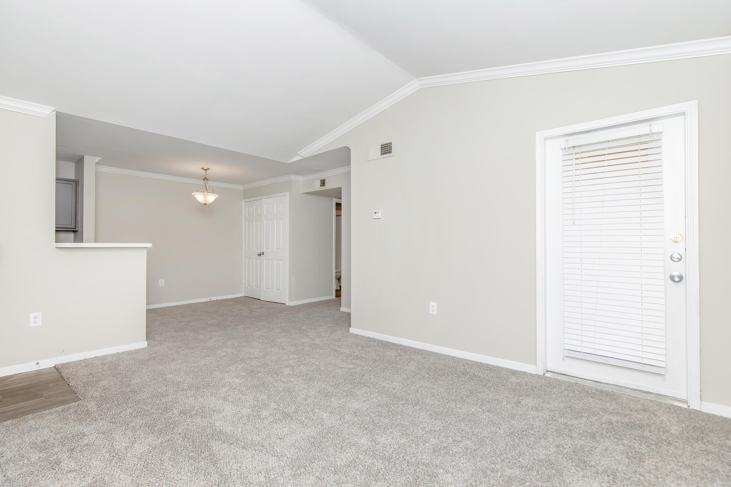 Spacious living area with light gray carpet, neutral walls, and a vaulted ceiling. A small dining area is visible to the left with a chandelier hanging above. To the right, a door with blinds leads outside. The overall design is simple and inviting, ideal for comfortable living.