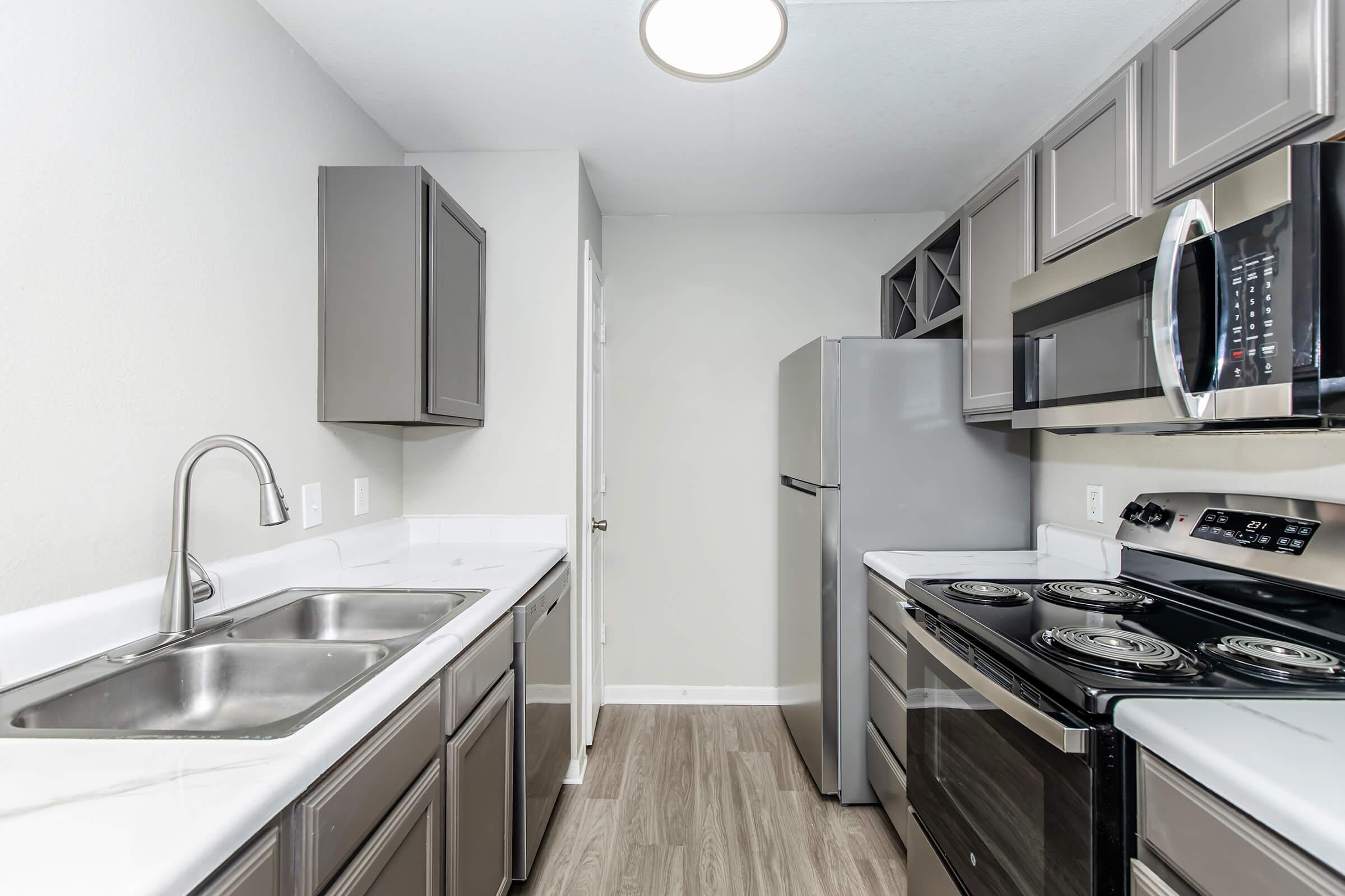 Modern kitchen featuring gray cabinets, a double sink, stainless steel appliances including a refrigerator and an oven, and a light fixture. The space has a neutral-colored wall and wooden flooring, creating a clean and inviting atmosphere.