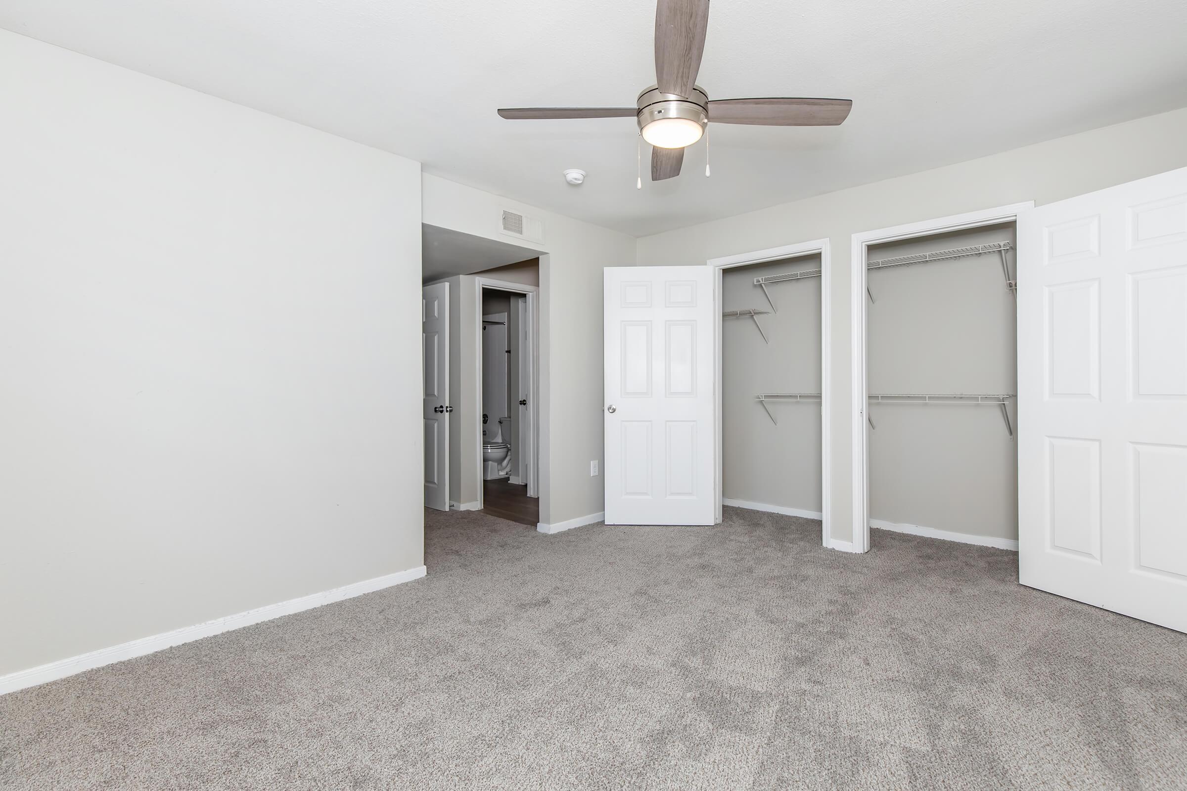 A spacious, empty bedroom with light gray carpet and white walls. A ceiling fan is installed, and there are two closets with open doors. One door leads to a bathroom area, partially visible. The room has a bright and airy feel, with natural light enhancing the neutral color palette.