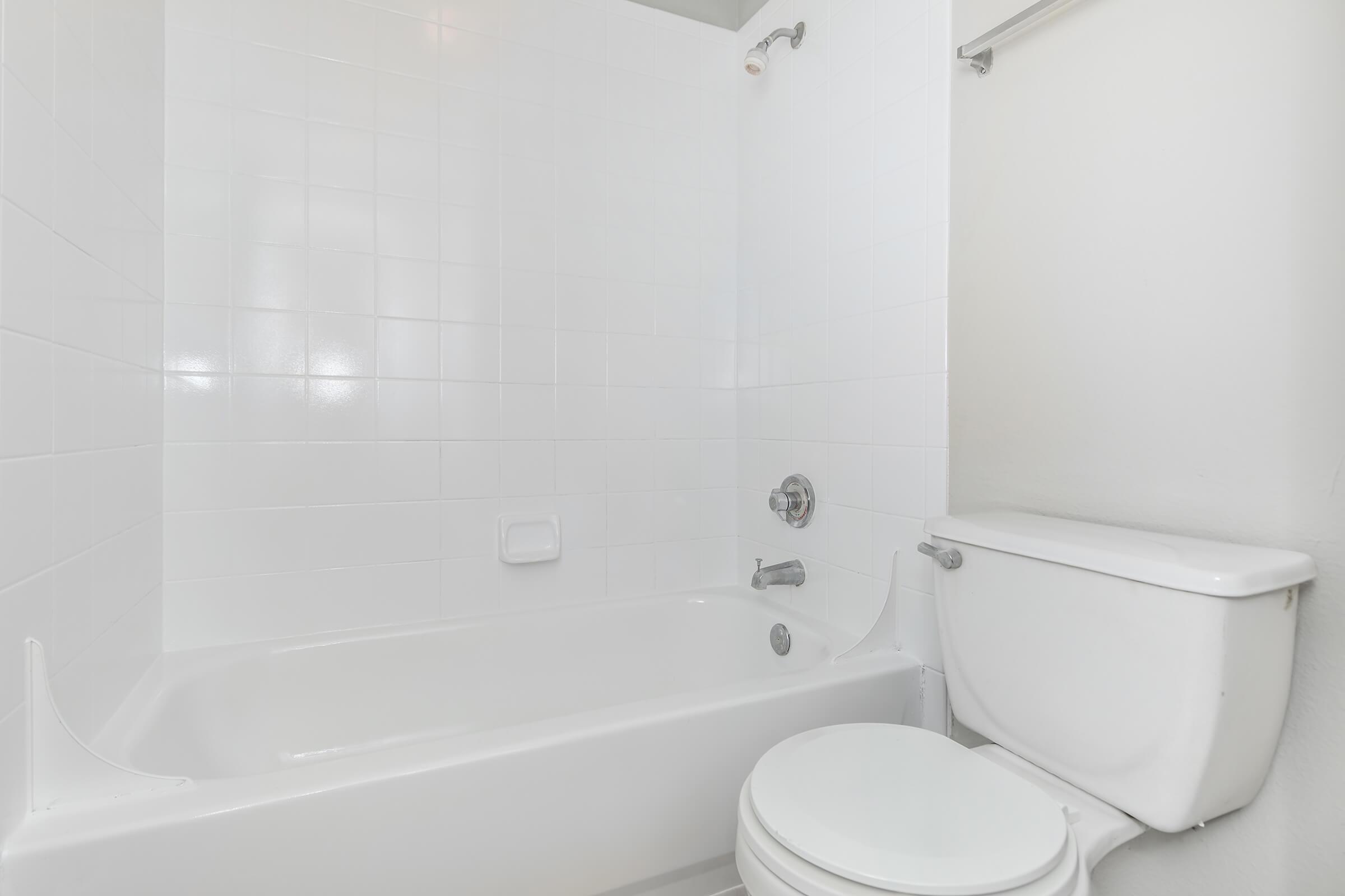 A clean, minimalist bathroom featuring a white bathtub with a showerhead, a white toilet, and tiled walls. The space is well-lit, emphasizing the bright, uncluttered design.