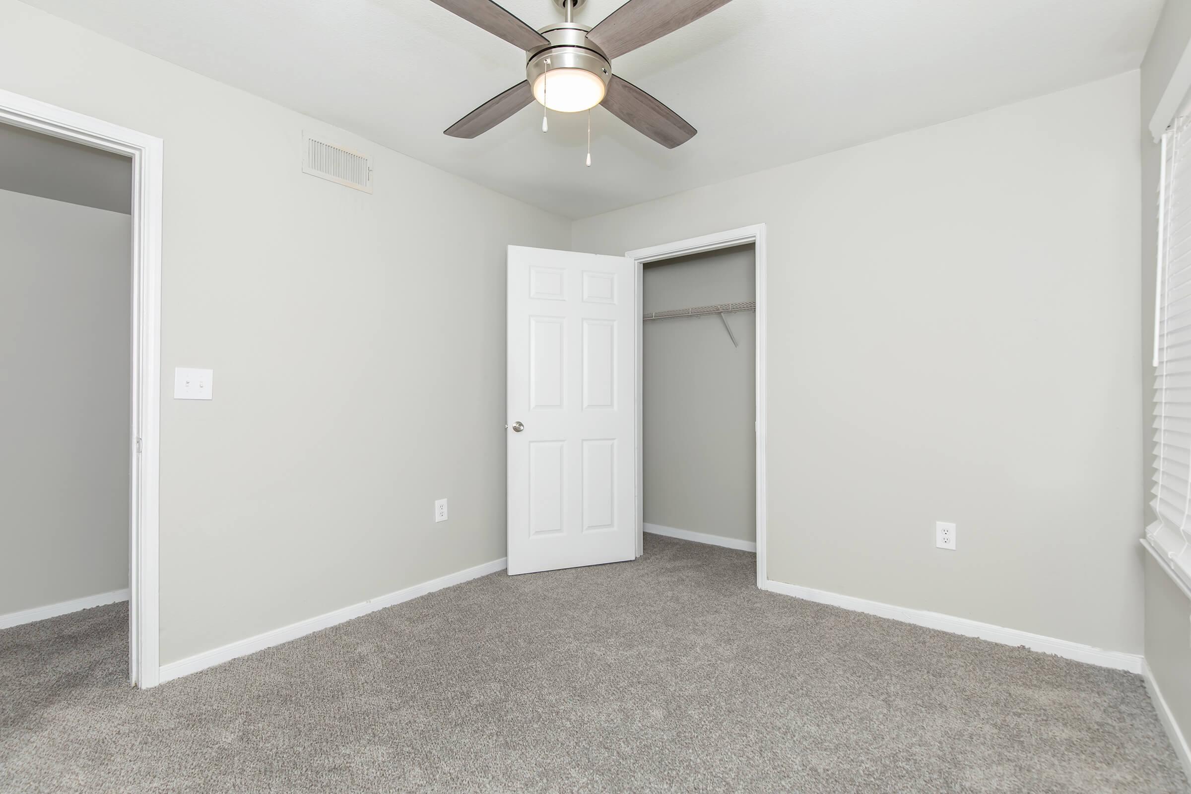A well-lit, empty bedroom featuring light gray walls and plush carpet. There is a ceiling fan with three blades, a closet with a sliding door on the left, and a white door leading to another room. Natural light enters through a window with blinds, enhancing the space's bright and clean appearance.