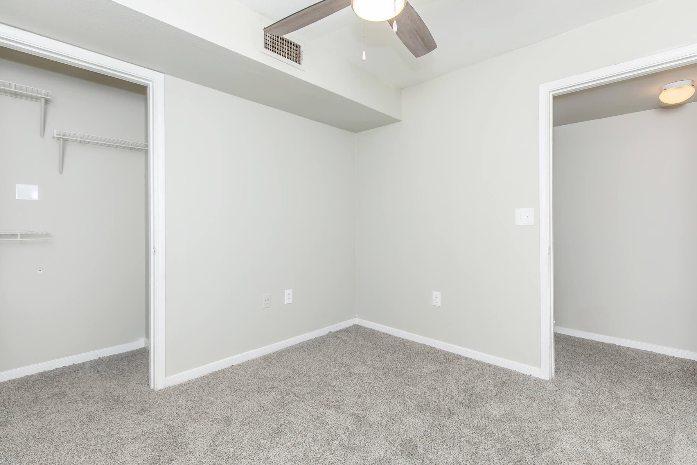 A clean, empty room with light gray walls and plush carpet. Two doorways lead to adjacent spaces, and a ceiling fan is visible overhead. The room has minimal decor, creating a spacious and open feel.