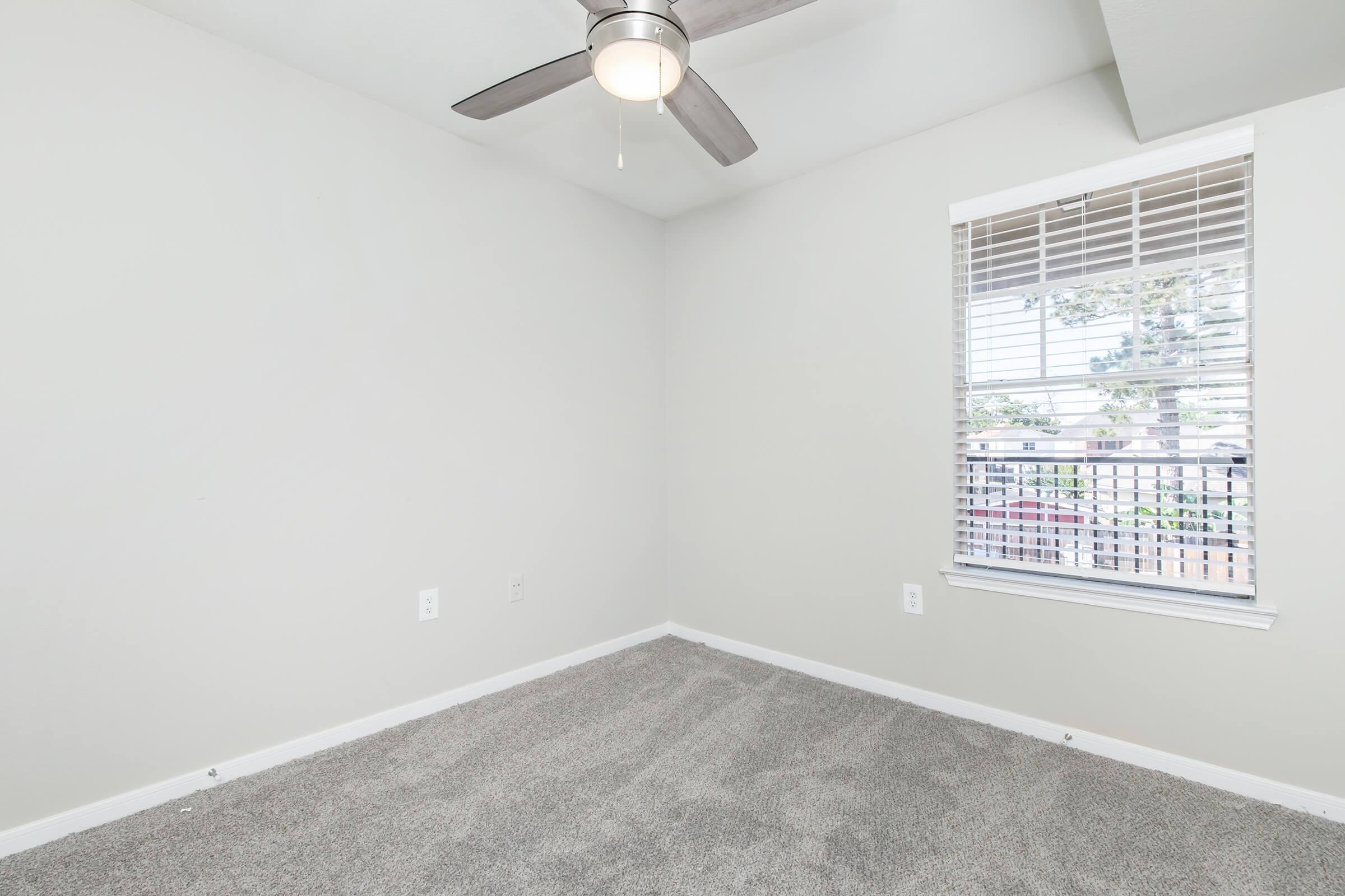 A light and airy room featuring grey carpet, a ceiling fan, and a window with blinds offering a view outside. The walls are painted in a soft neutral color, creating a serene atmosphere. No furniture is present, highlighting the spaciousness of the area.