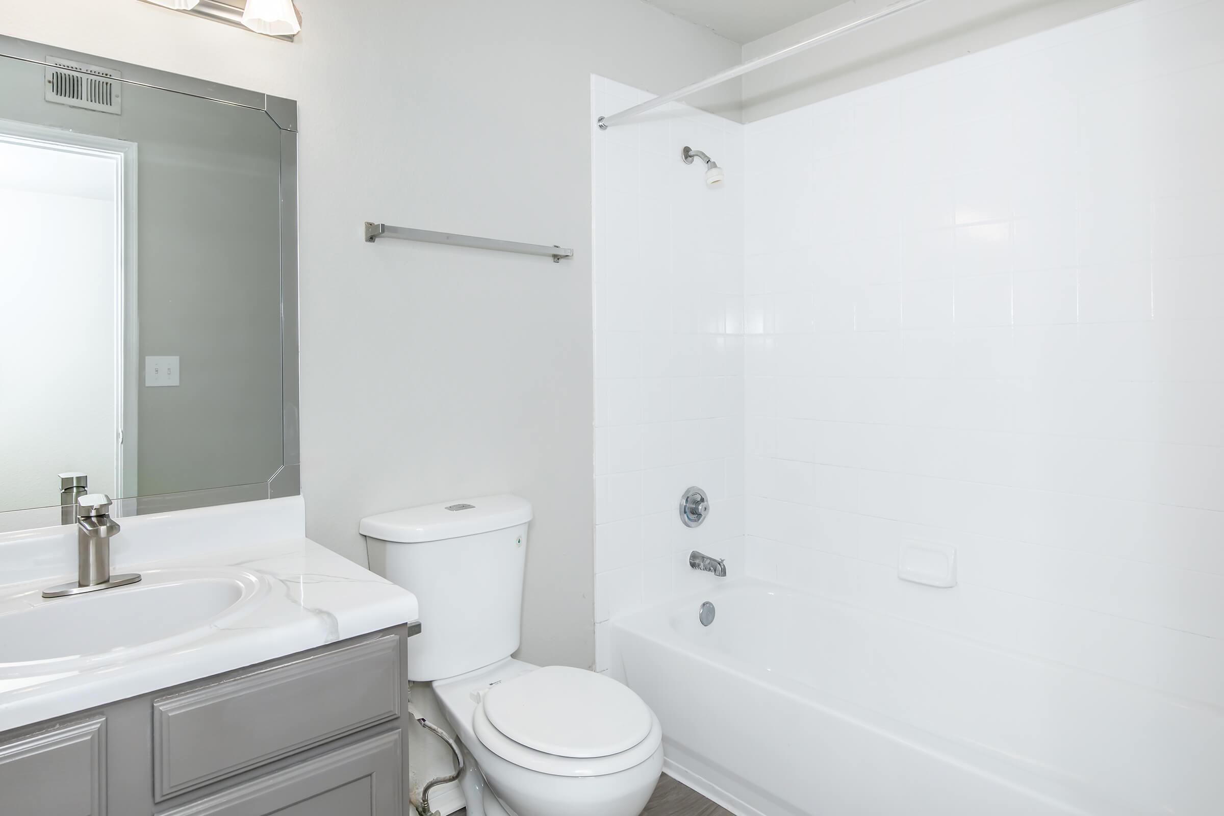 A clean and bright bathroom featuring a white bathtub with a shower, a modern toilet, a single sink with a mirror above it, and a gray cabinet. The walls are painted light, creating an airy atmosphere, and there is a towel bar installed on the wall.