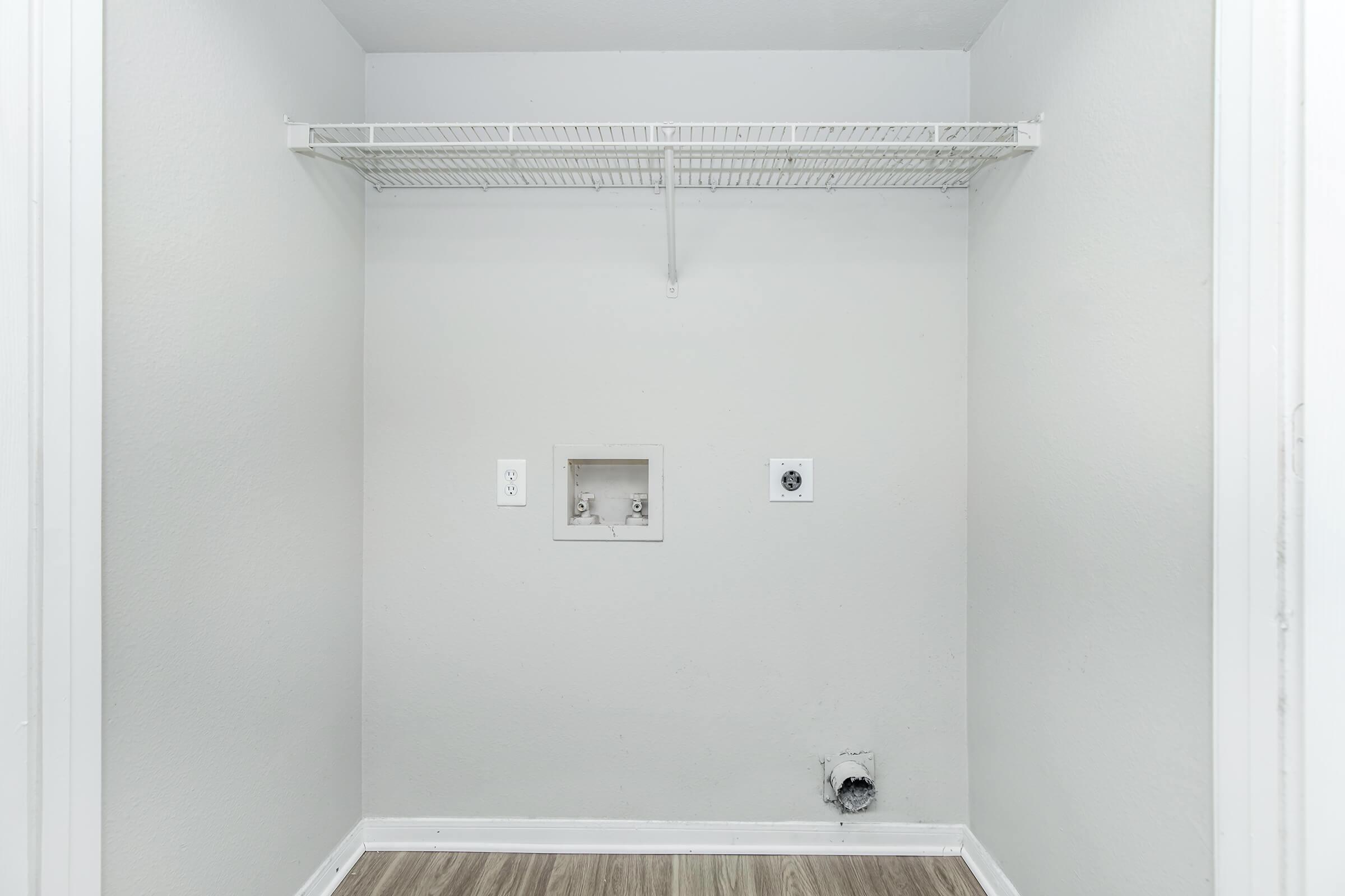 An empty room with light gray walls and a wooden floor. There is a metal wire shelf mounted on the wall and a small opening on one side, likely for appliances. No furniture or decorations are present, creating a minimalistic appearance.