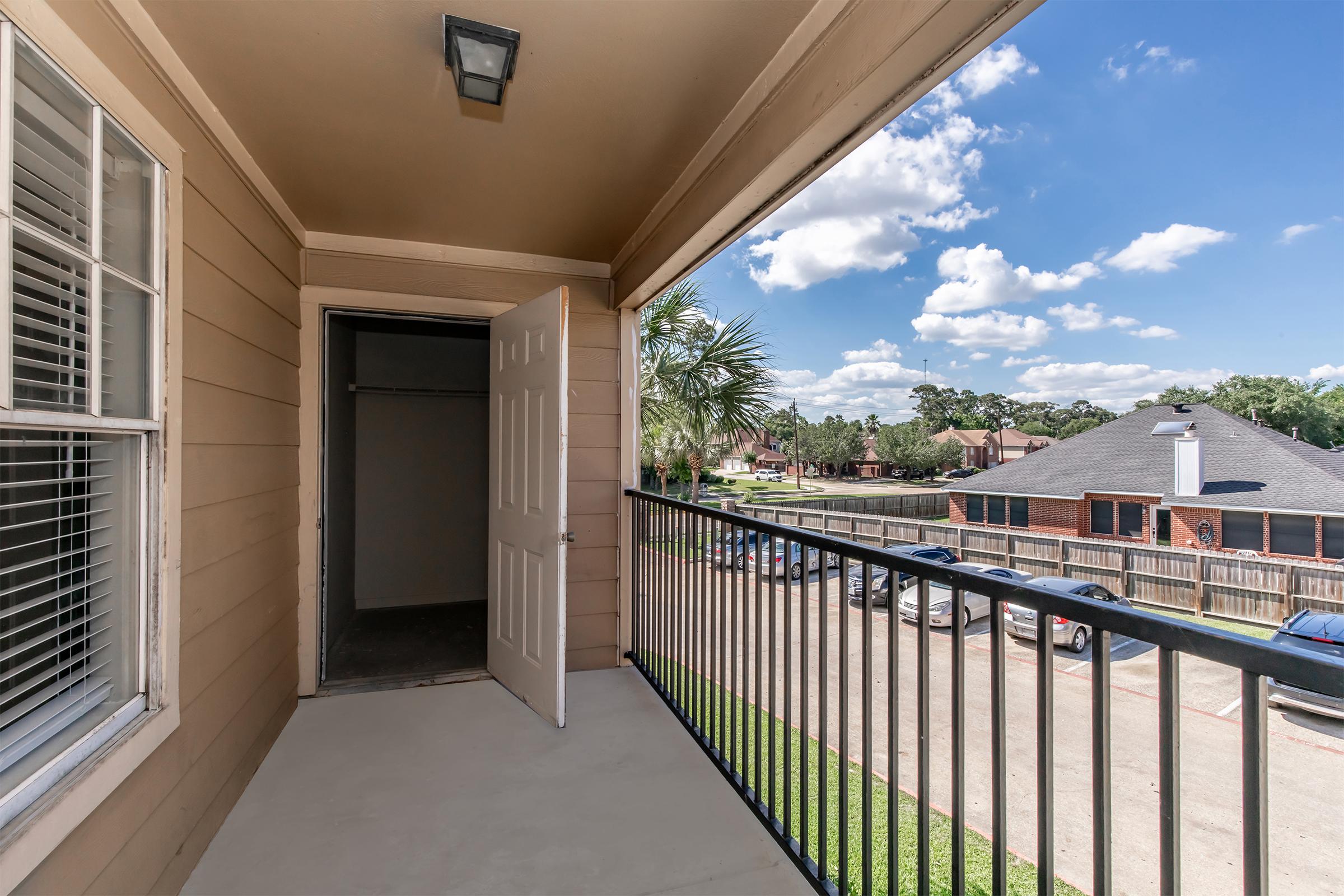 A view from a balcony featuring an open door leading into a room. The balcony has a railing with a clear view of a neighborhood, including trees and buildings under a partly cloudy sky. The scene conveys a sense of openness and outdoor space.