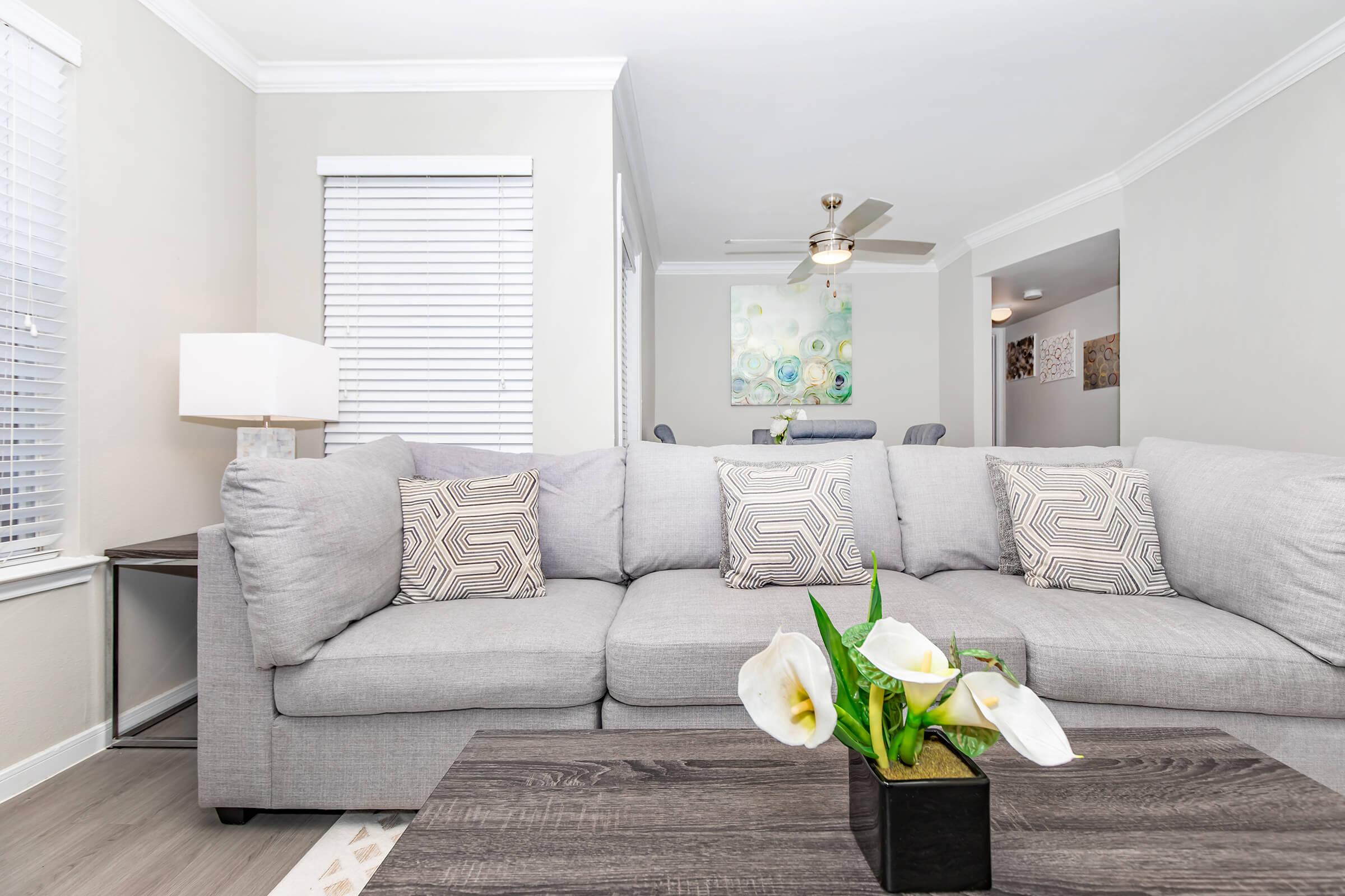 A modern living room featuring a gray sofa with patterned cushions, a dark wooden coffee table adorned with white flowers, and a lamp. Behind the sofa, there are windows with white blinds, and a glimpse of a dining area decorated with abstract art can be seen in the background.