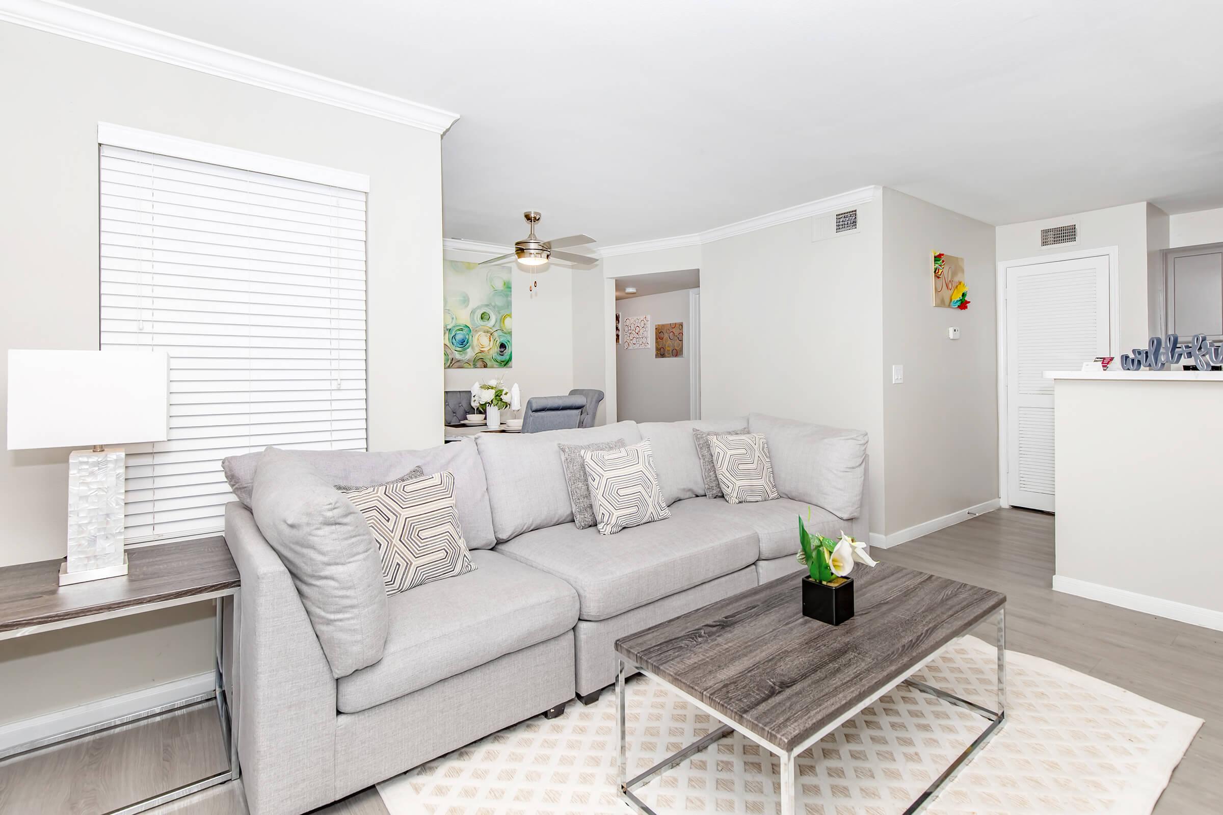 A modern living room featuring a light gray sectional sofa adorned with patterned pillows, a sleek coffee table, and a decorative plant. The walls are painted in soft gray tones, and a ceiling fan is visible. Natural light filters through a window with white blinds, creating a cozy atmosphere.