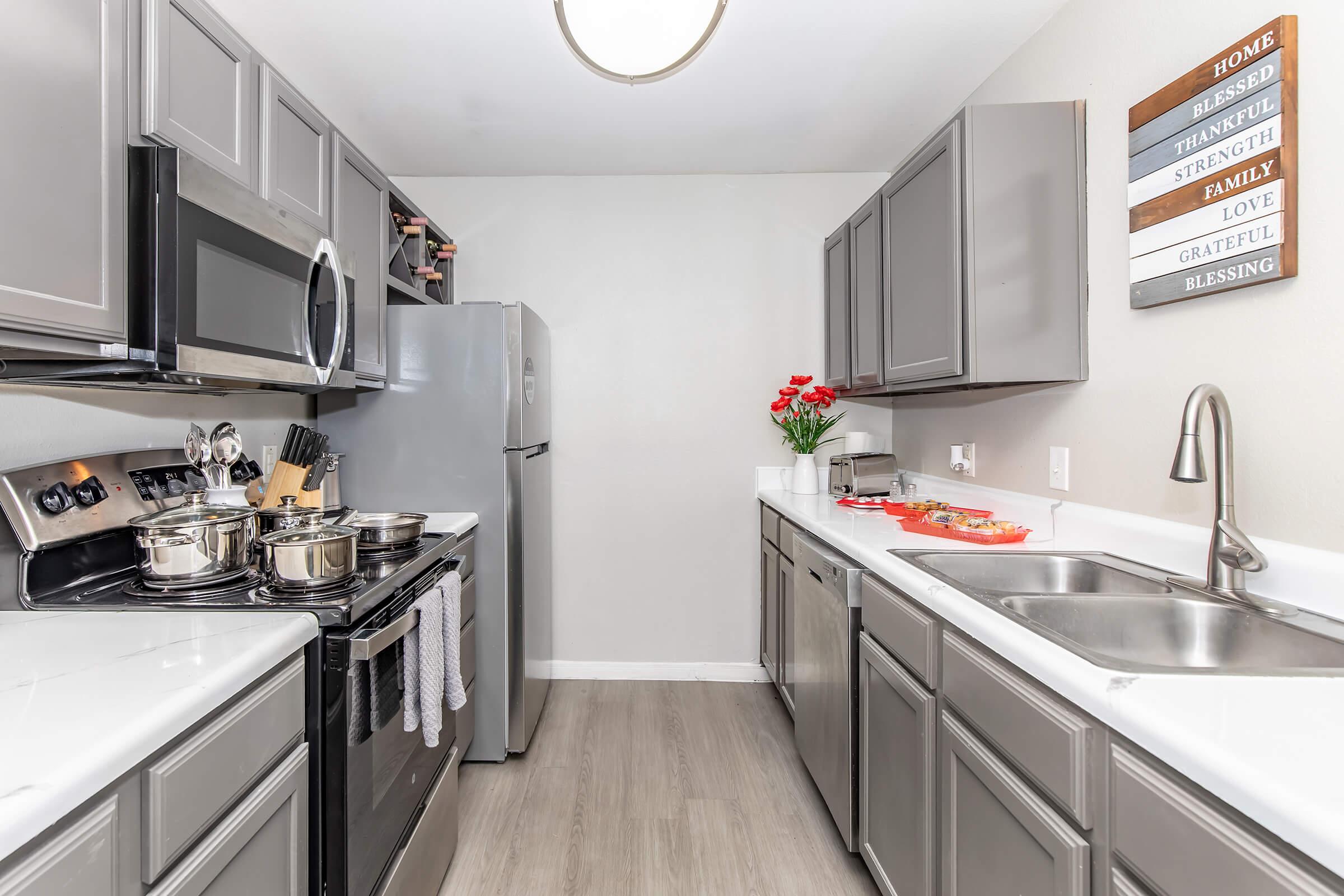 Modern kitchen featuring gray cabinetry and stainless steel appliances. There are pots on the stove, a refrigerator, and a double sink. Red flowers in a vase add a pop of color. Wall decor includes a wooden sign with words like "Home," "Family," and "Blessing." Bright and inviting atmosphere.