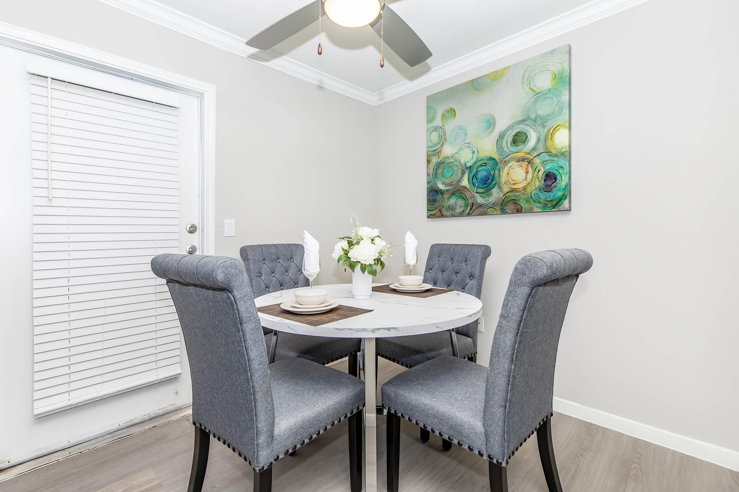 A modern dining area featuring a round marble dining table surrounded by four upholstered gray chairs. On the wall, there's a colorful abstract painting with circular patterns. A ceiling fan is visible above, and a closed white window with horizontal blinds is on the left. Fresh flowers are arranged in the center of the table.