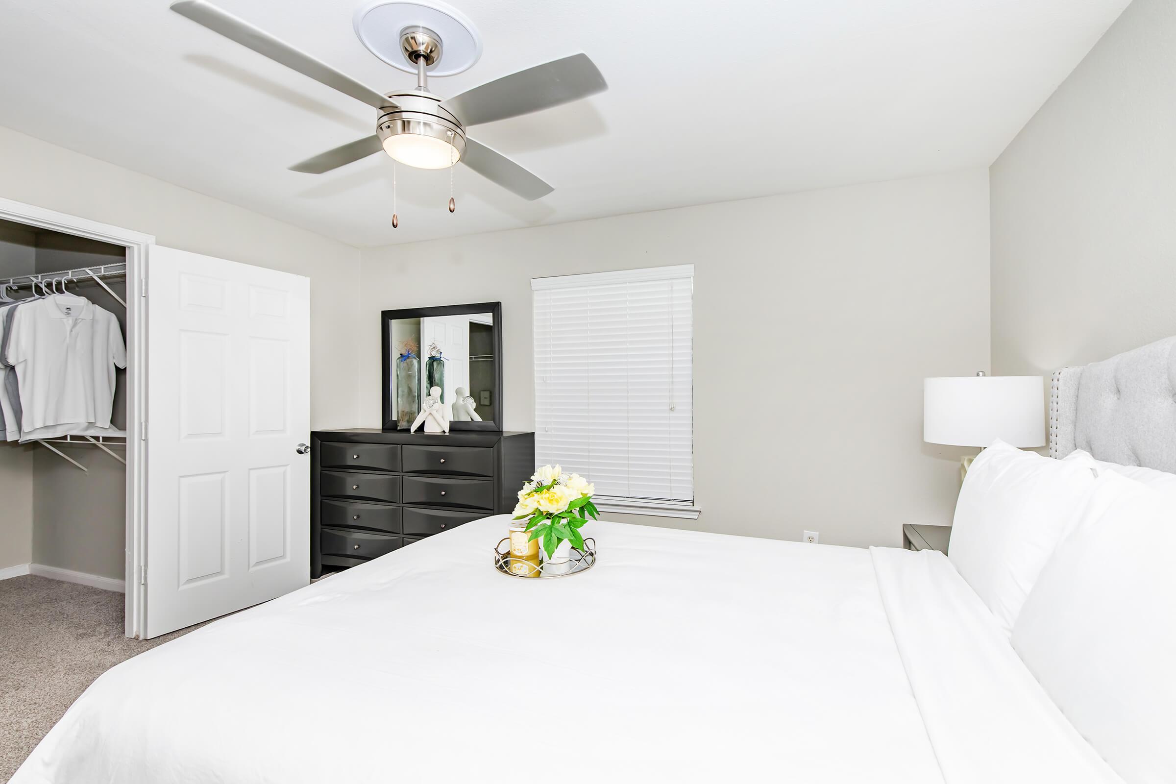 A cozy bedroom featuring a neatly made bed with white linens and a decorative tray with flowers. A ceiling fan hangs overhead, and there is a dresser with multiple drawers. A closet with hanging clothes is visible, along with a closed white door and a window with blinds, allowing natural light to enter.