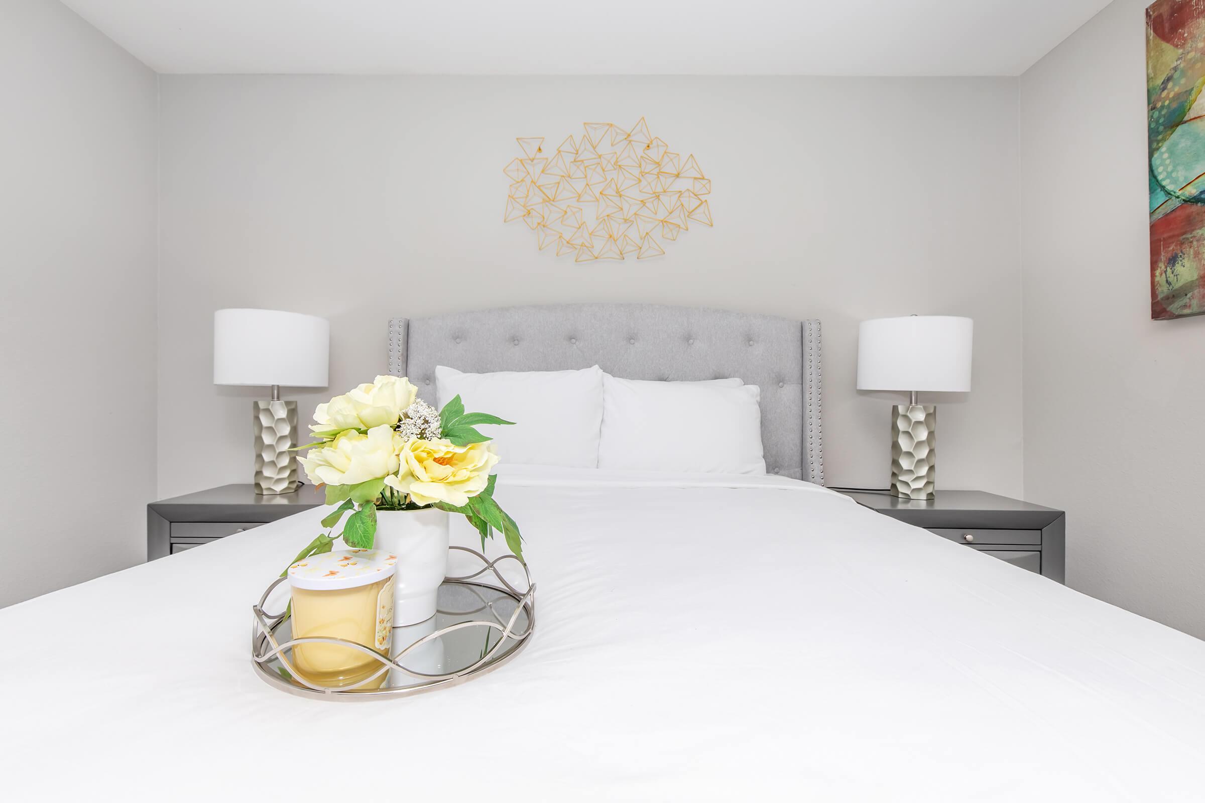 A neatly made bed with white linens and gray upholstered headboard, flanked by stylish lamps on nightstands. A tray holds a candle and a vase with yellow and white flowers, adding a warm touch to the modern decor. A geometric wall art piece complements the serene color scheme.