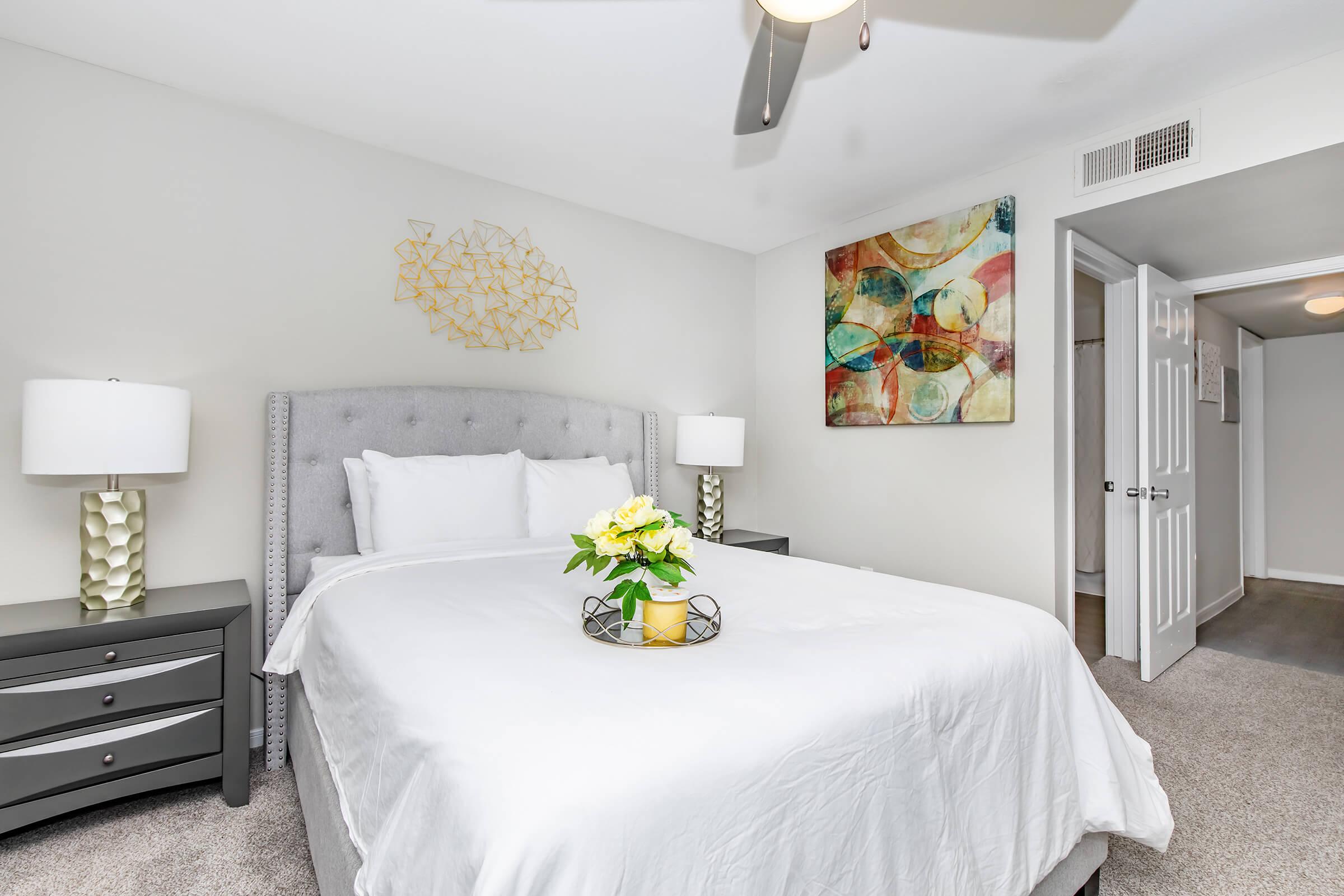 A modern bedroom featuring a light gray upholstered bed with white bedding, two stylish nightstands with lamps, and a decorative piece on the wall. A vase of yellow flowers sits on a circular tray on the bed. The room has neutral-colored walls and a soft carpet, creating a cozy atmosphere.