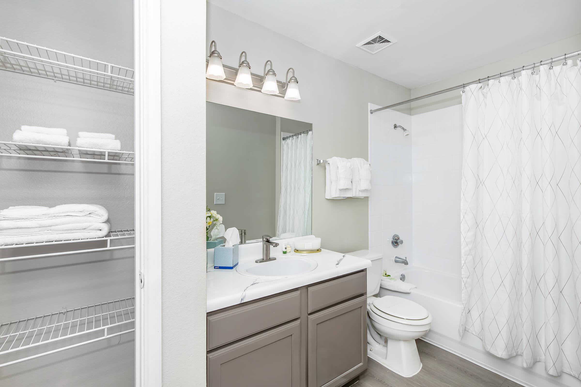 A modern bathroom featuring a white bathtub with a shower curtain, a clean vanity with a sink, and a large mirror above it. There are neatly stacked white towels on a shelf nearby. The color scheme is neutral with gray walls and wooden flooring, creating a bright and spacious feel.