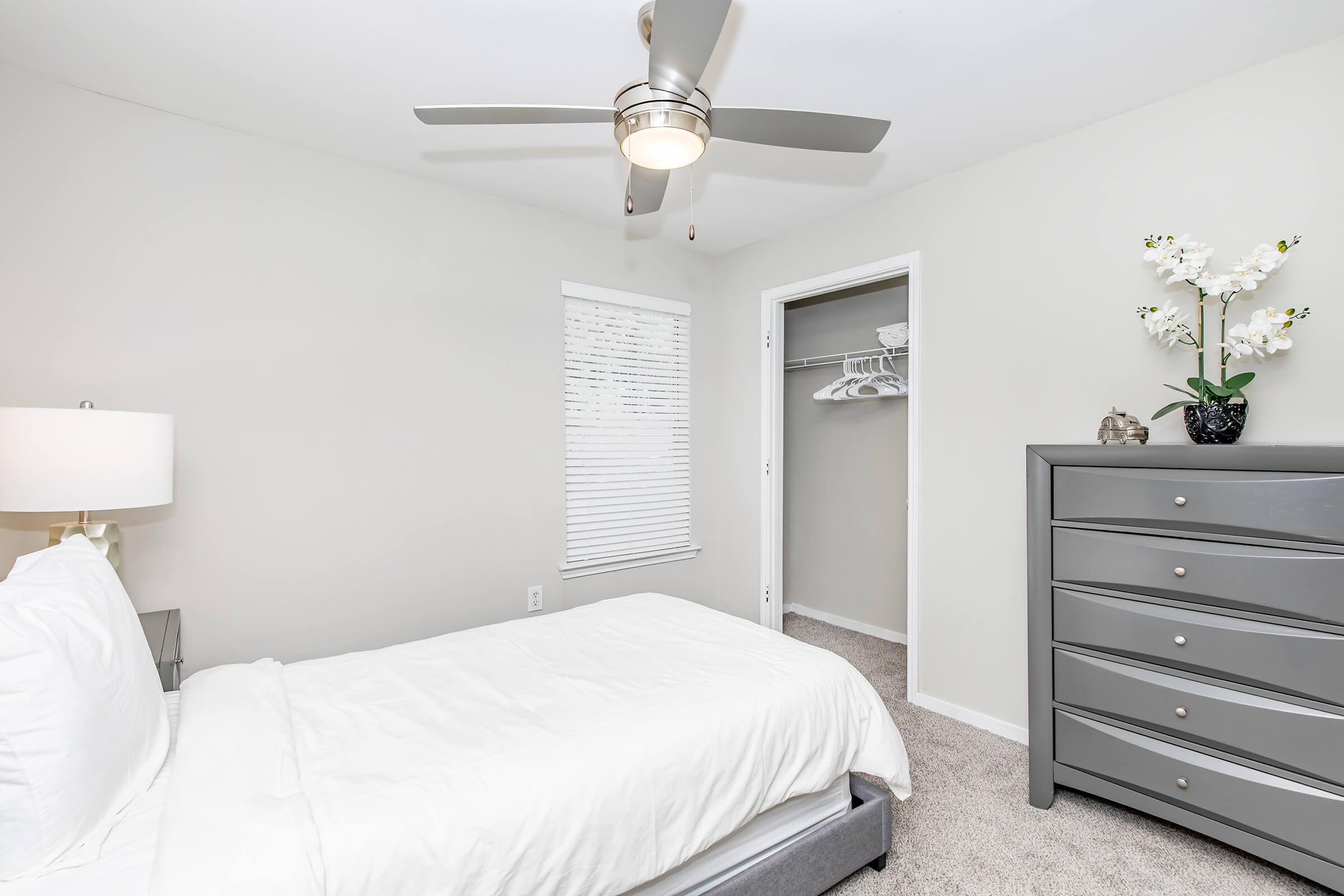 A cozy bedroom featuring a neatly made bed with a white comforter, a light gray dresser with multiple drawers, and a ceiling fan. There is a window with white blinds allowing natural light in and a closet with hangers visible. A small plant with white flowers adds a touch of decor to the room.