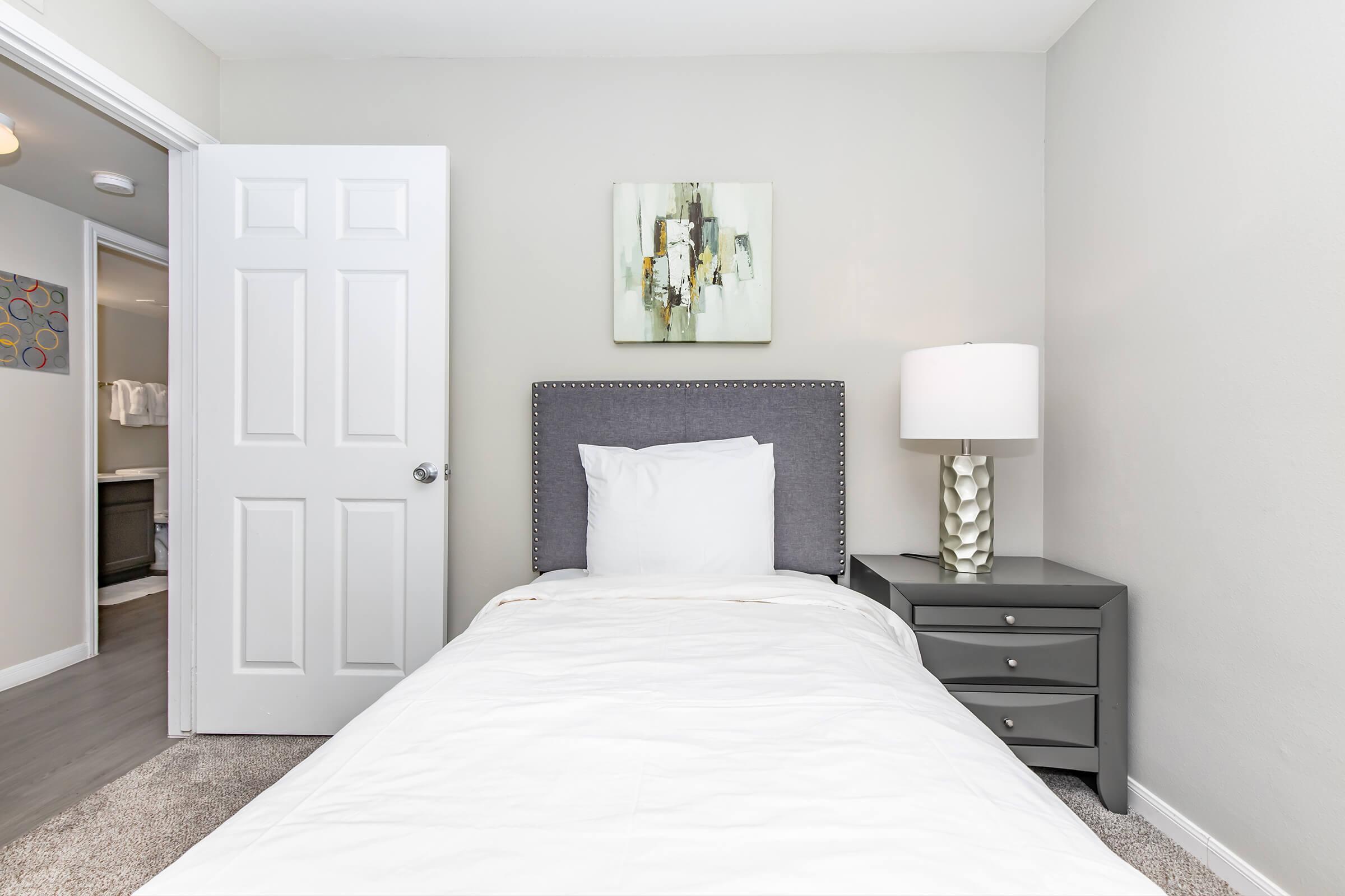 Cozy bedroom featuring a single bed with white bedding, a modern bedside table, and a decorative lamp. A piece of abstract wall art hangs above the bed, and a door in the background leads to another room. The walls are painted light gray, creating a bright and inviting atmosphere.