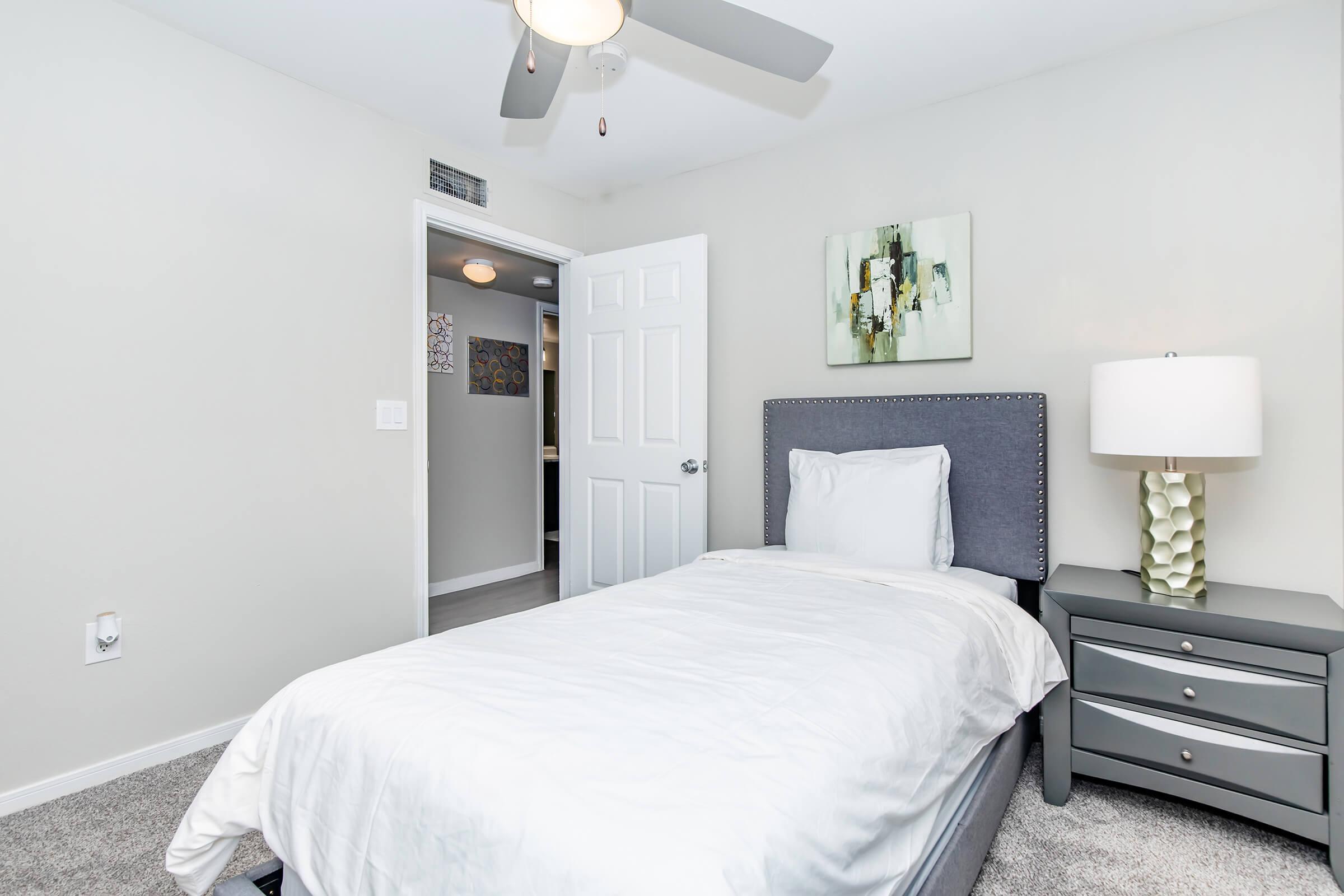 A cozy bedroom featuring a single bed with white linens, a gray upholstered headboard, and a nightstand with a lamp. The room has light-colored walls and carpet, with a piece of abstract wall art. A door leads to a bathroom, and a ceiling fan is installed above.