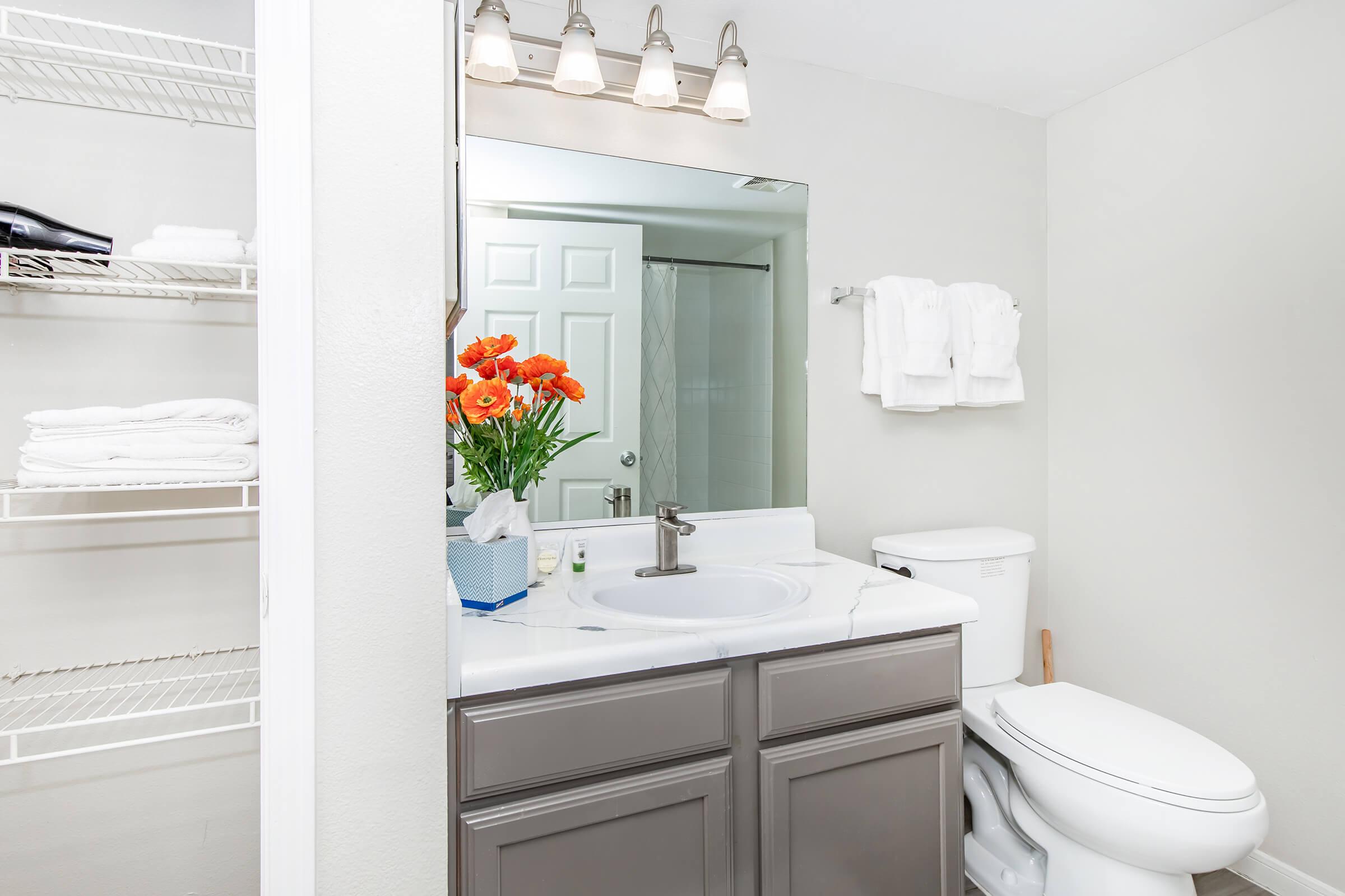 A bright and clean bathroom featuring a vanity with a sink and a mirror above. There are neatly stacked white towels on a rack and a bouquet of orange flowers on the counter. A shower area is visible in the background, alongside a white toilet. The walls are painted a light gray.