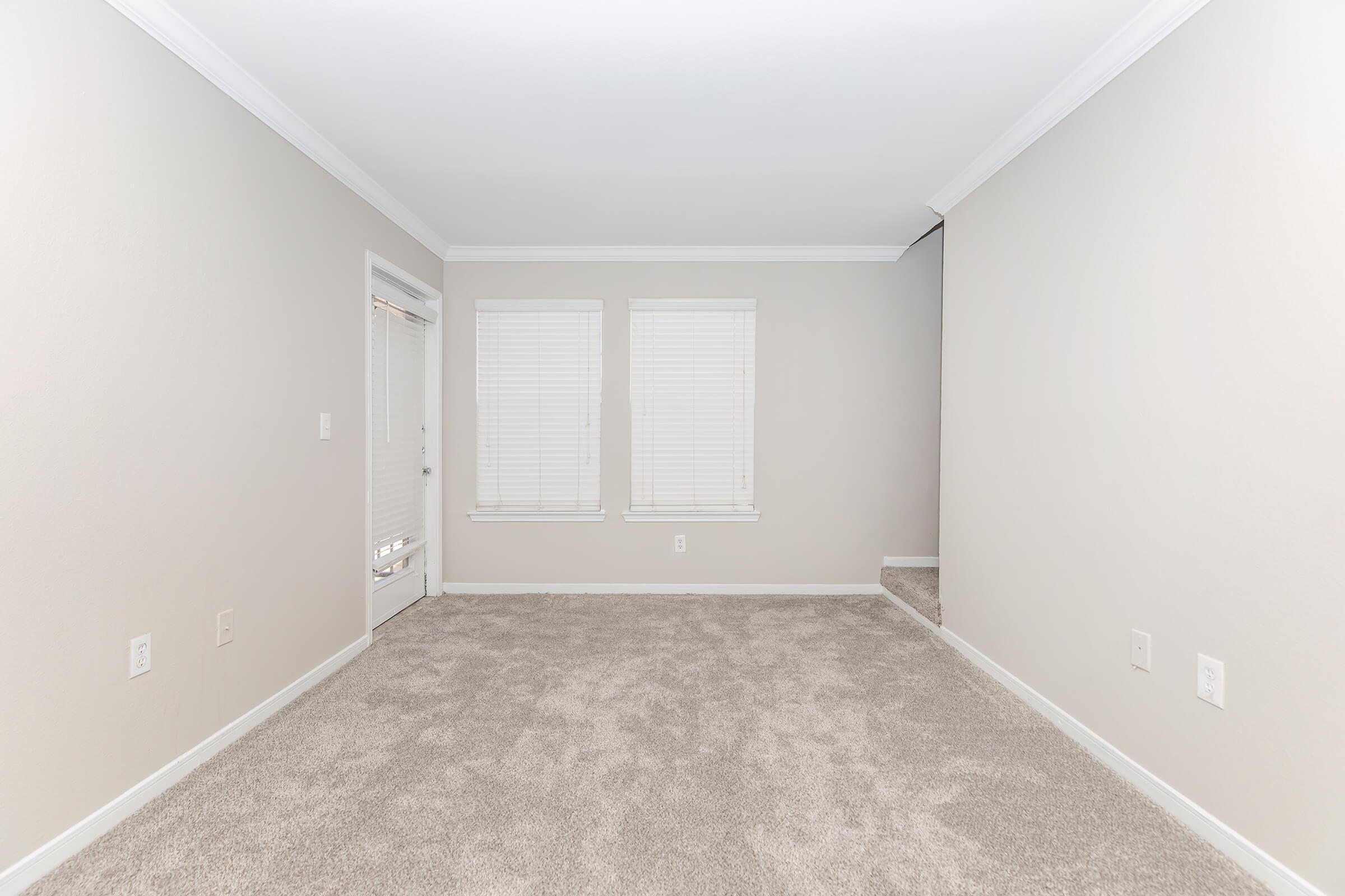 A spacious, empty room with light gray walls and plush carpet. Two narrow windows with white blinds allow natural light. A door on the left leads outside, and there is a staircase on the right leading to another area. The overall decor is minimalistic and modern.