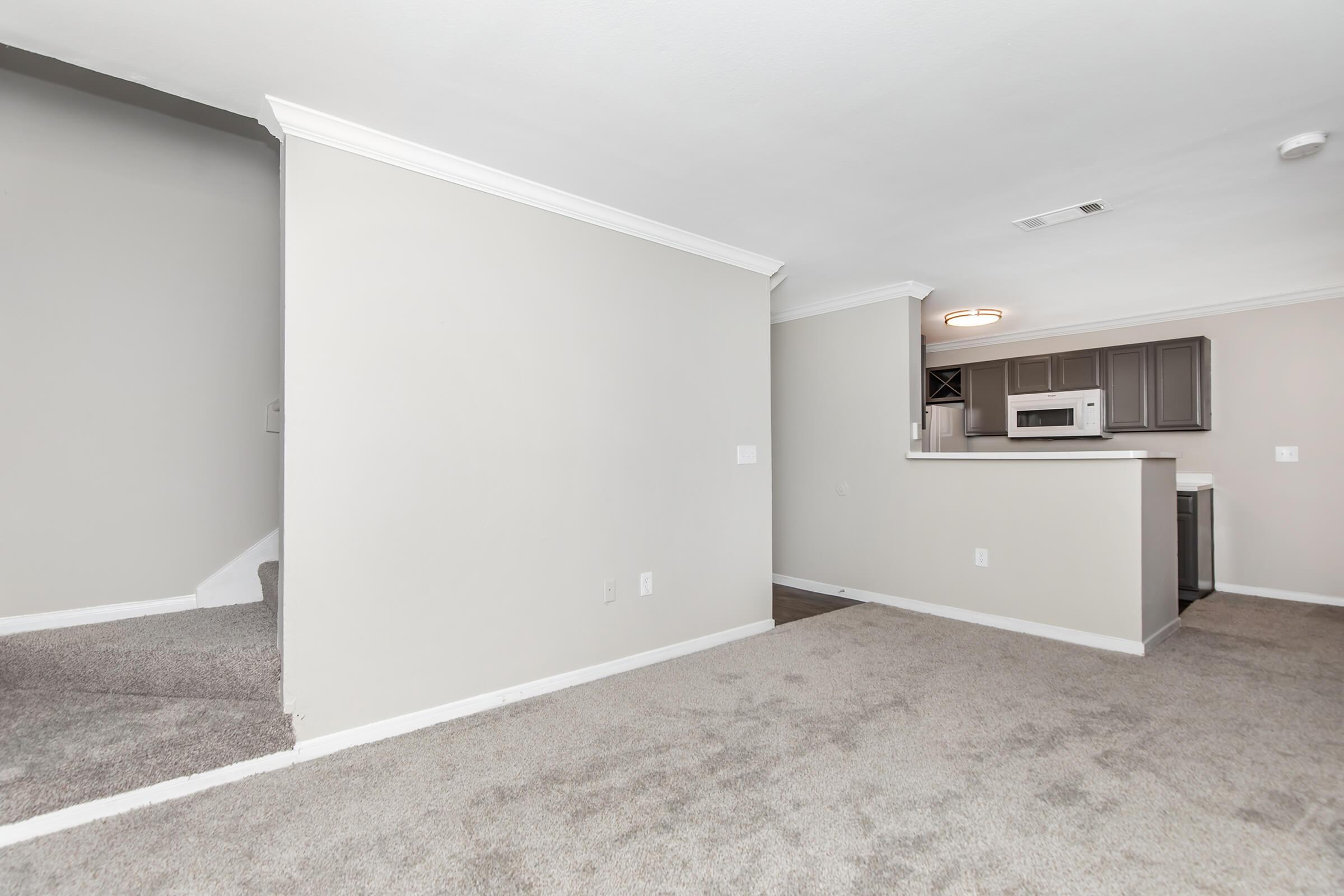 A spacious living area with light-colored walls and carpeted flooring. To the left, there is a staircase leading to another level, while the kitchen area is partially visible in the background, featuring modern cabinets and appliances. The room is well-lit and invites a cozy atmosphere.