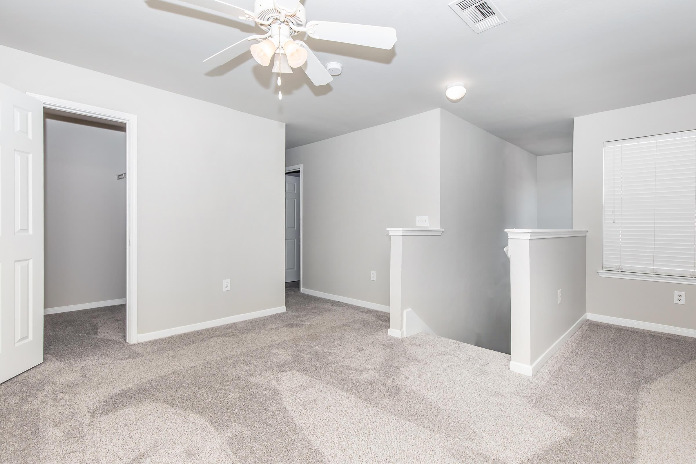 A spacious, well-lit corner of a room featuring light gray walls and plush, beige carpet. A ceiling fan is visible above, and there's a doorway on the left leading to a closet. The room has an open feel with another door on the right and a window allowing natural light to enter.