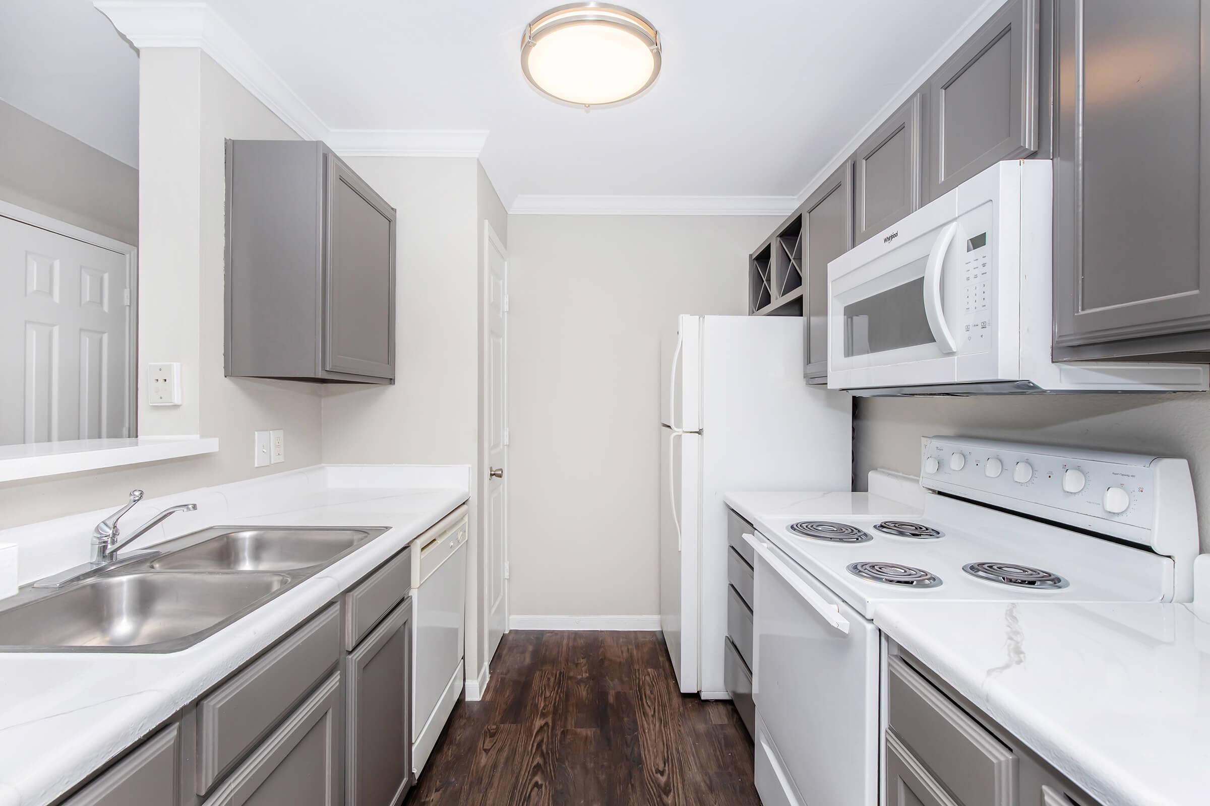 A modern kitchen featuring gray cabinetry and white appliances. The layout includes a double sink, a refrigerator, a microwave, and an oven with a stovetop. The walls are painted light gray, and there's a light fixture on the ceiling. Dark wooden flooring contrasts with the bright kitchen elements.