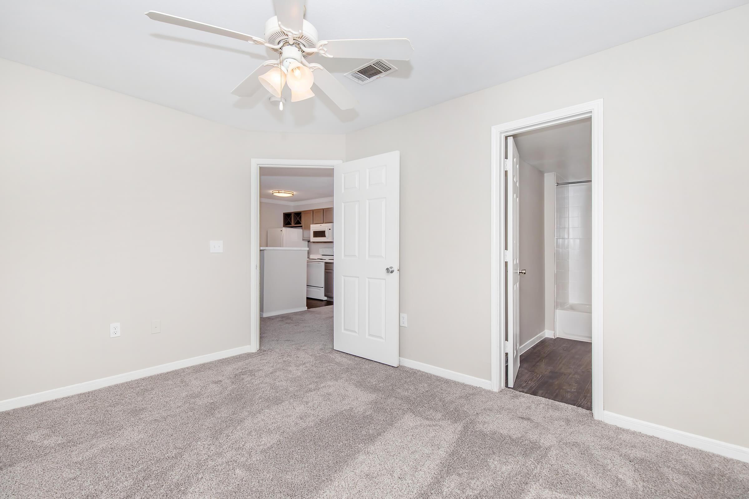 A spacious, light-colored bedroom with plush carpet flooring. The room features a ceiling fan and has open doors leading to a kitchen area and a bathroom. Natural light fills the space, creating a bright and inviting atmosphere. Neutral wall colors enhance the room's airy feel.