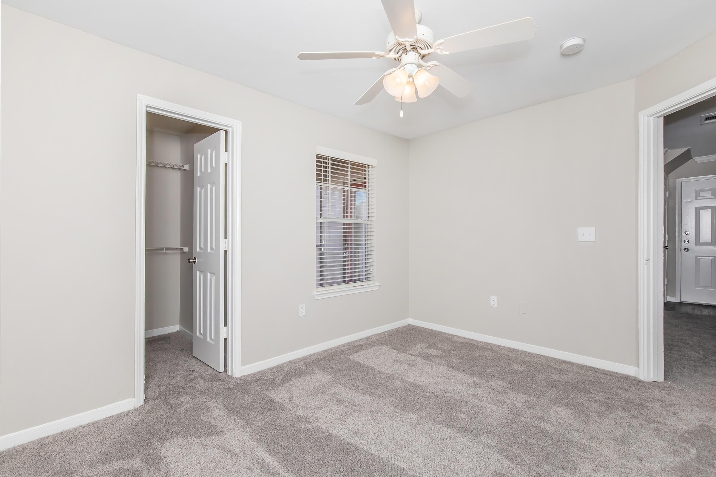Light and airy room featuring freshly painted beige walls, plush gray carpet, a ceiling fan, and a window with blinds. An empty closet is visible in one corner, while a door leads to another room. The overall atmosphere is clean and inviting, perfect for personalizing with furnishings.