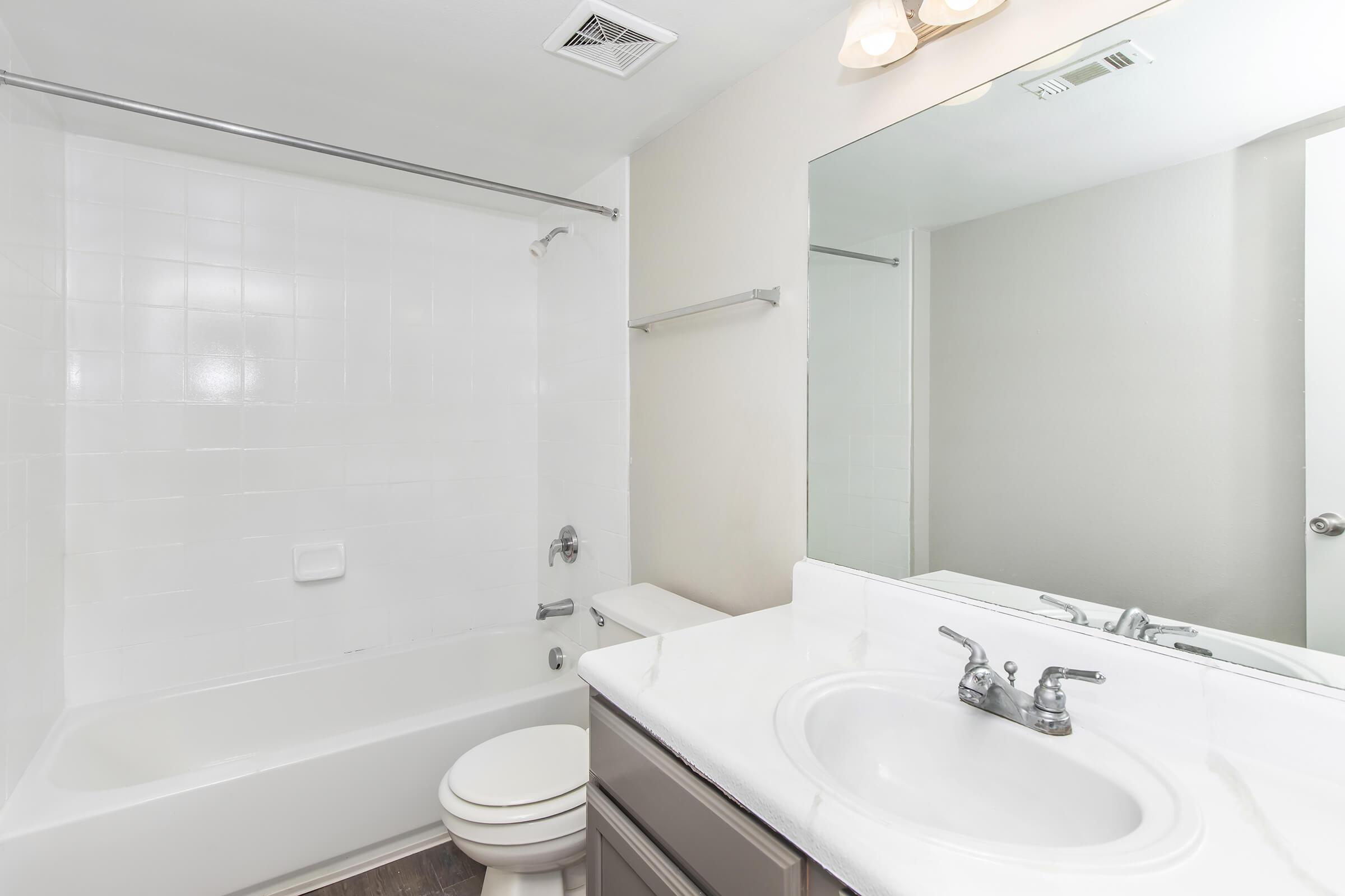 A clean, modern bathroom featuring a white tub with a shower, a white sink with a chrome faucet, a large mirror above the sink, and light-colored walls. There is also a toilet and a towel bar. The overall design is simple and functional, with good lighting.