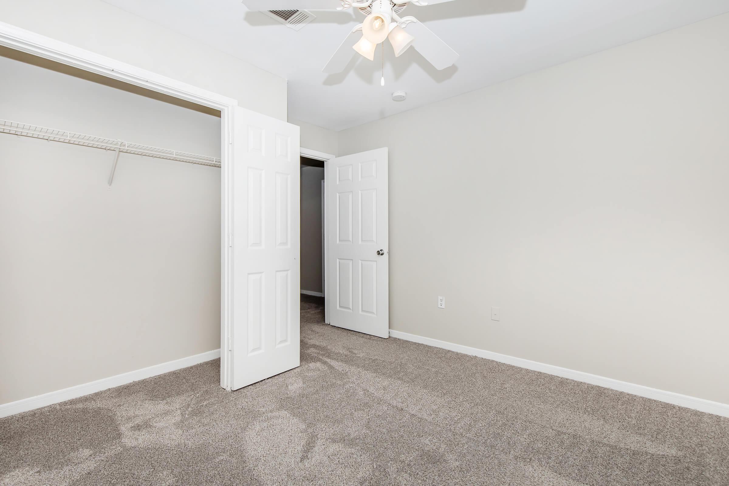 An empty room with light beige walls and light gray carpet. Two white double doors are open, leading to a closet on the left and another space beyond the right door. A ceiling fan is visible above. The room is well-lit, creating a bright and spacious appearance.