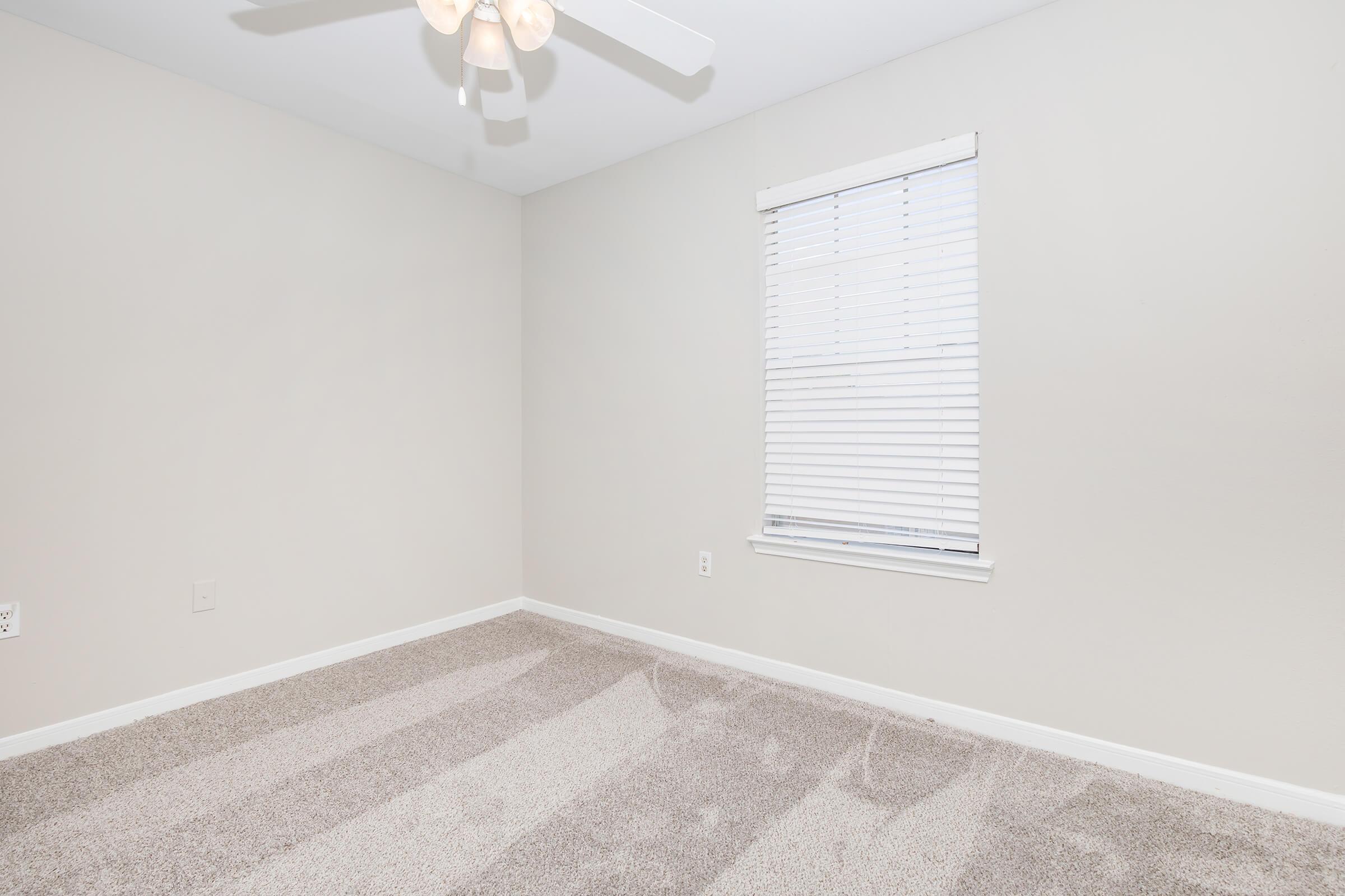 Empty room with light beige walls, a ceiling fan with light fixture, and a window with white blinds. The floor is covered in light tan carpet, and there are no furniture or decorations present. The room appears bright and spacious.