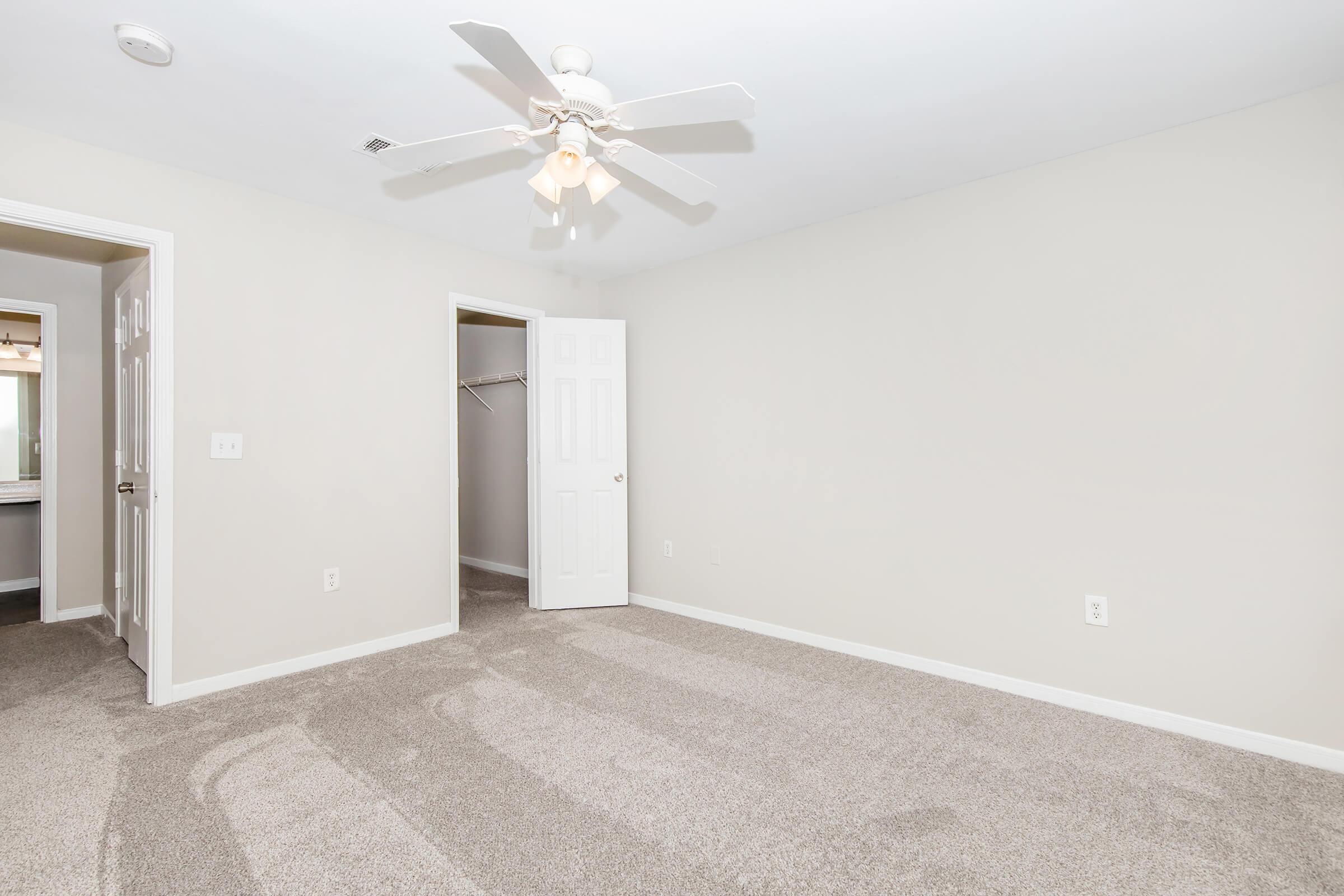A minimalistic bedroom featuring light-colored walls and soft carpet. The room includes a ceiling fan, an open closet with no doors, and a doorway leading to a bathroom. Natural light brightens the space, creating an inviting atmosphere.