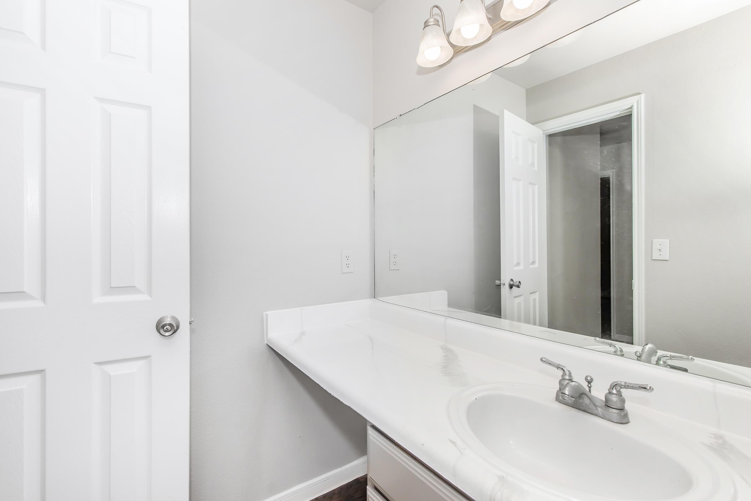 A clean, modern bathroom featuring a white countertop with a sink, a large mirror above, and a light fixture with multiple bulbs. A white door is partially visible on the left, and another door is seen in the background. The walls are painted in a light color, contributing to a spacious feel.