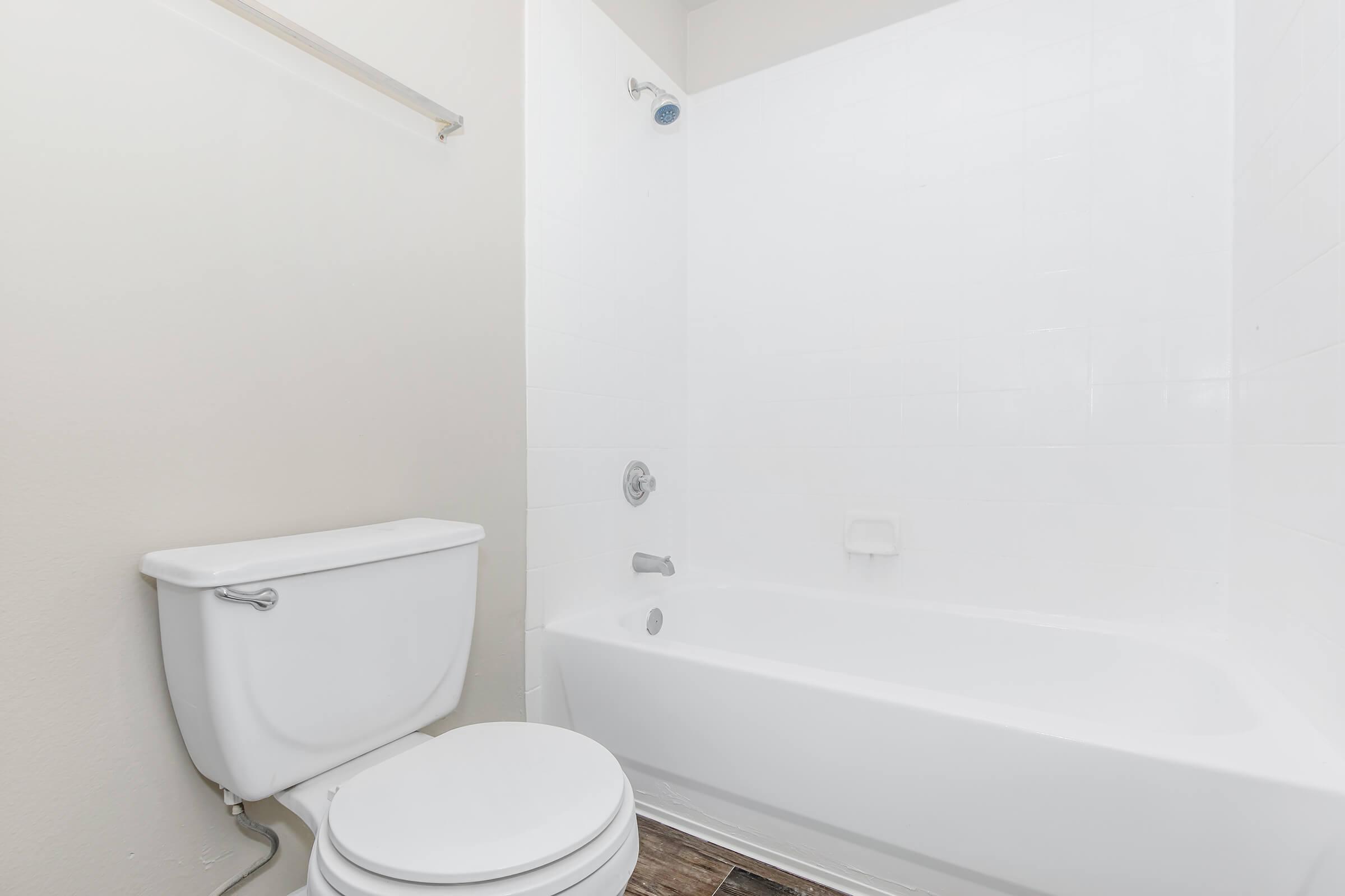 A clean, minimalist bathroom featuring a white bathtub, a white toilet, and light-colored walls. The bathroom has a showerhead mounted above the tub and a towel bar on the wall, with wooden flooring partially visible. The space is well-lit and tidy, emphasizing a simple design.