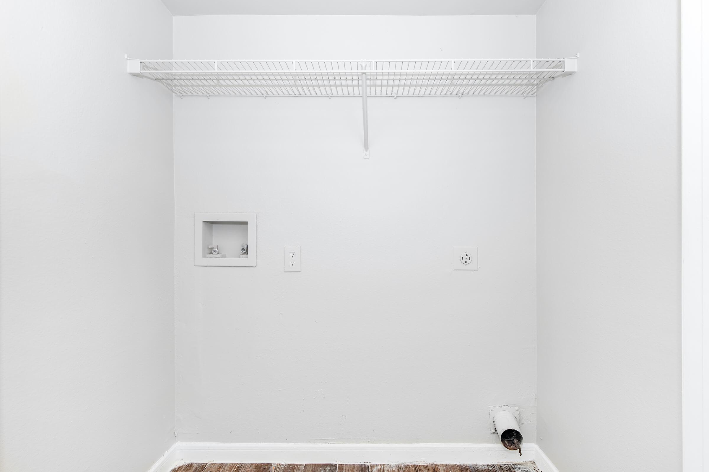 A small, empty closet with white walls and a wire shelf. There are two electrical outlets and a small opening on one wall, possibly for a vent or other utility. The floor is bare, likely hardwood, and there’s no visible storage or items inside. The overall space is bright and uncluttered.