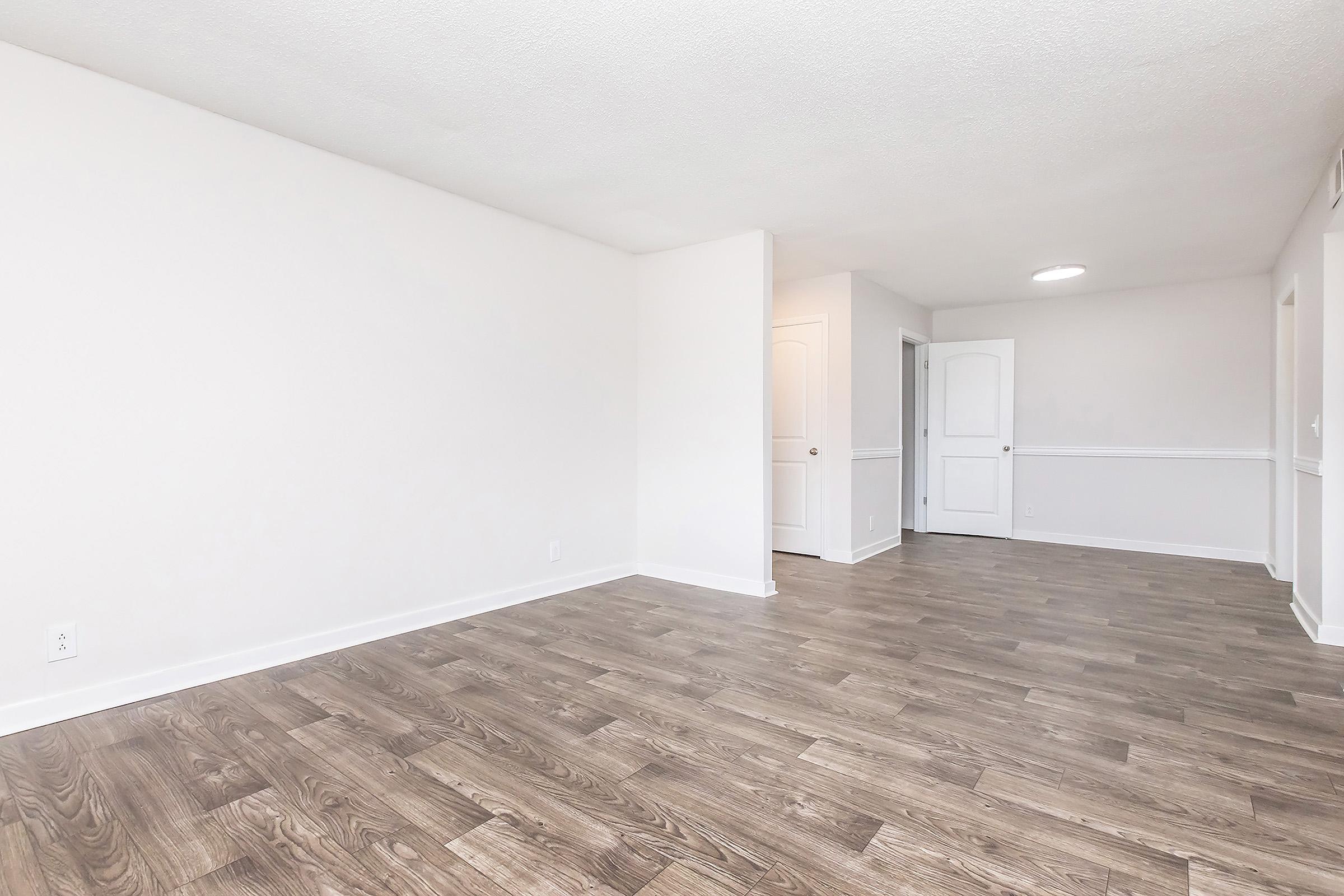 A spacious, empty room with light-colored walls and modern wooden flooring. The room features a doorway leading to another area, and there's a ceiling light fixture visible. The overall atmosphere is bright and inviting, suitable for various purposes like living or office space.