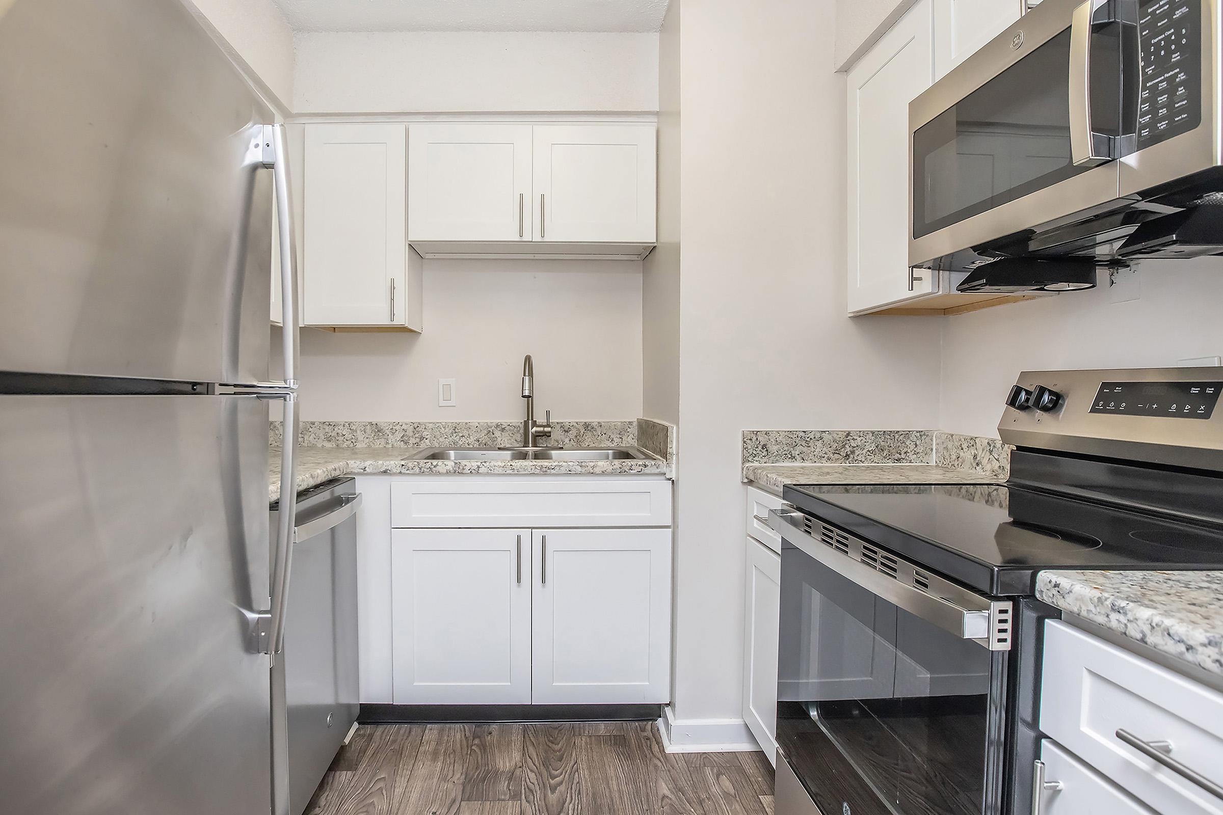 Modern kitchen featuring stainless steel appliances, including a refrigerator, microwave, and oven. The space has white cabinetry and granite countertops, with a sink visible between the cabinets. The flooring is a dark wood finish, creating a stylish contrast with the lighter elements.