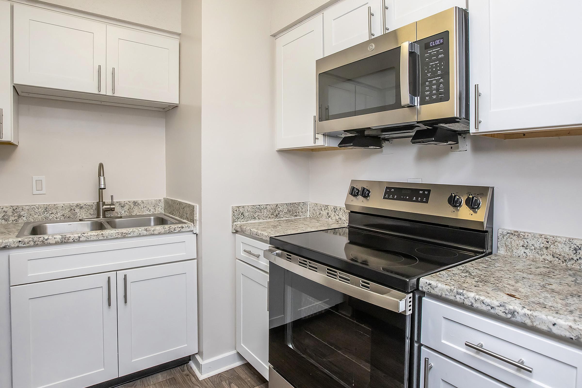 Modern kitchen featuring white cabinetry, granite countertops, stainless steel microwave, and an electric range. The sink is located on one side, with overhead cabinets providing storage. The flooring is a light wood finish, creating a bright and functional cooking space.