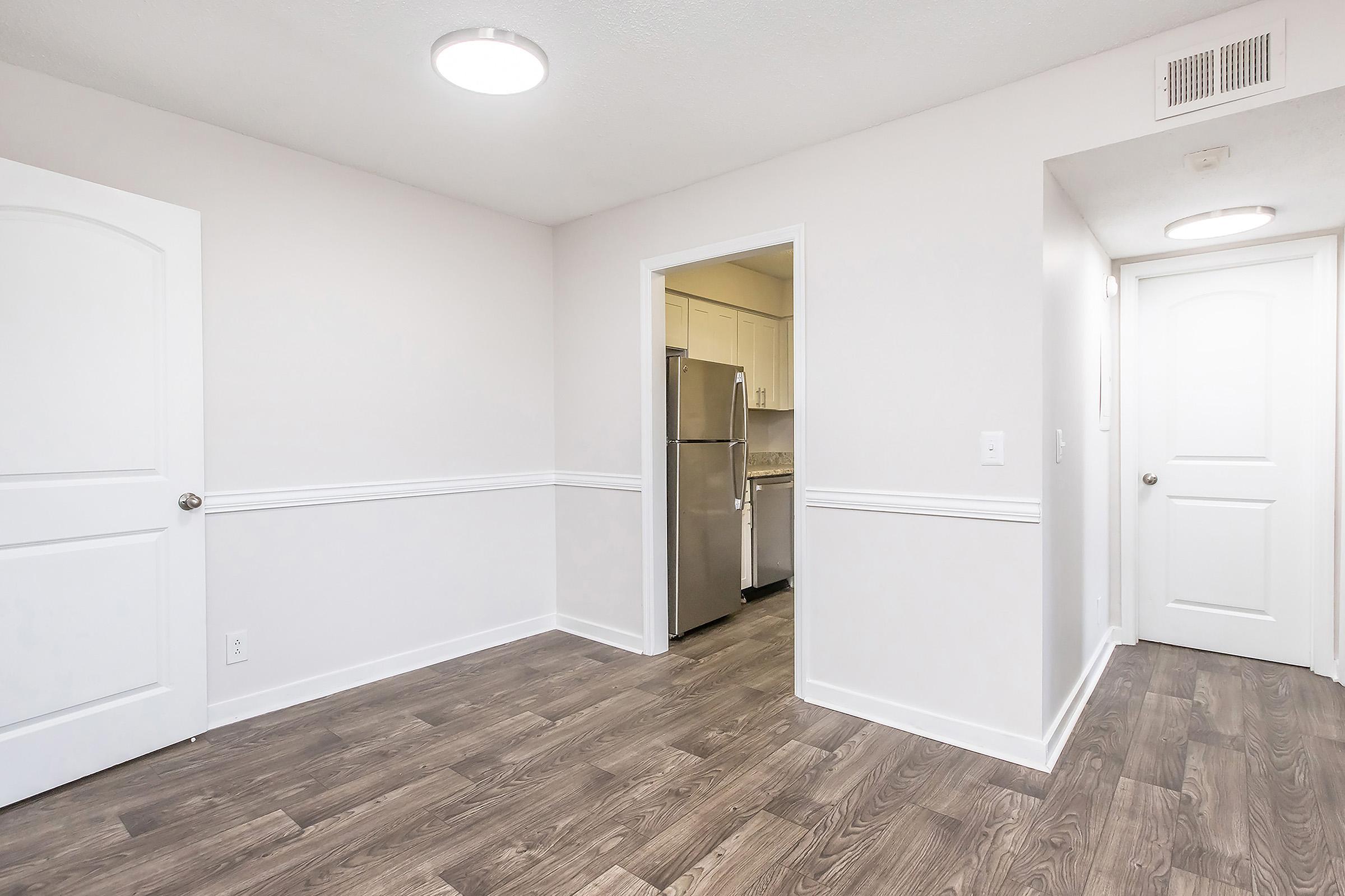 An interior view of a modern apartment with light-colored walls and laminate flooring. There's a doorway leading to a kitchen visible in the background with stainless steel appliances. Two doors are seen on the left and right, and ceiling lights provide illumination in the space.