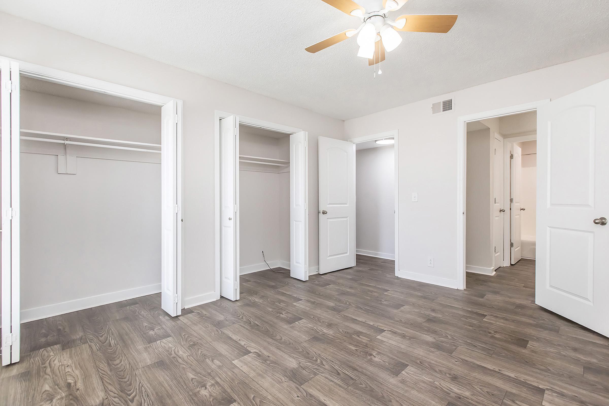 A bright, spacious bedroom featuring light-colored walls, a ceiling fan, and wood-like flooring. Two open closets are visible on the left, while a doorway leads to a bathroom on the right. The overall space is well-lit and clean, with a neutral decor perfect for various styles.