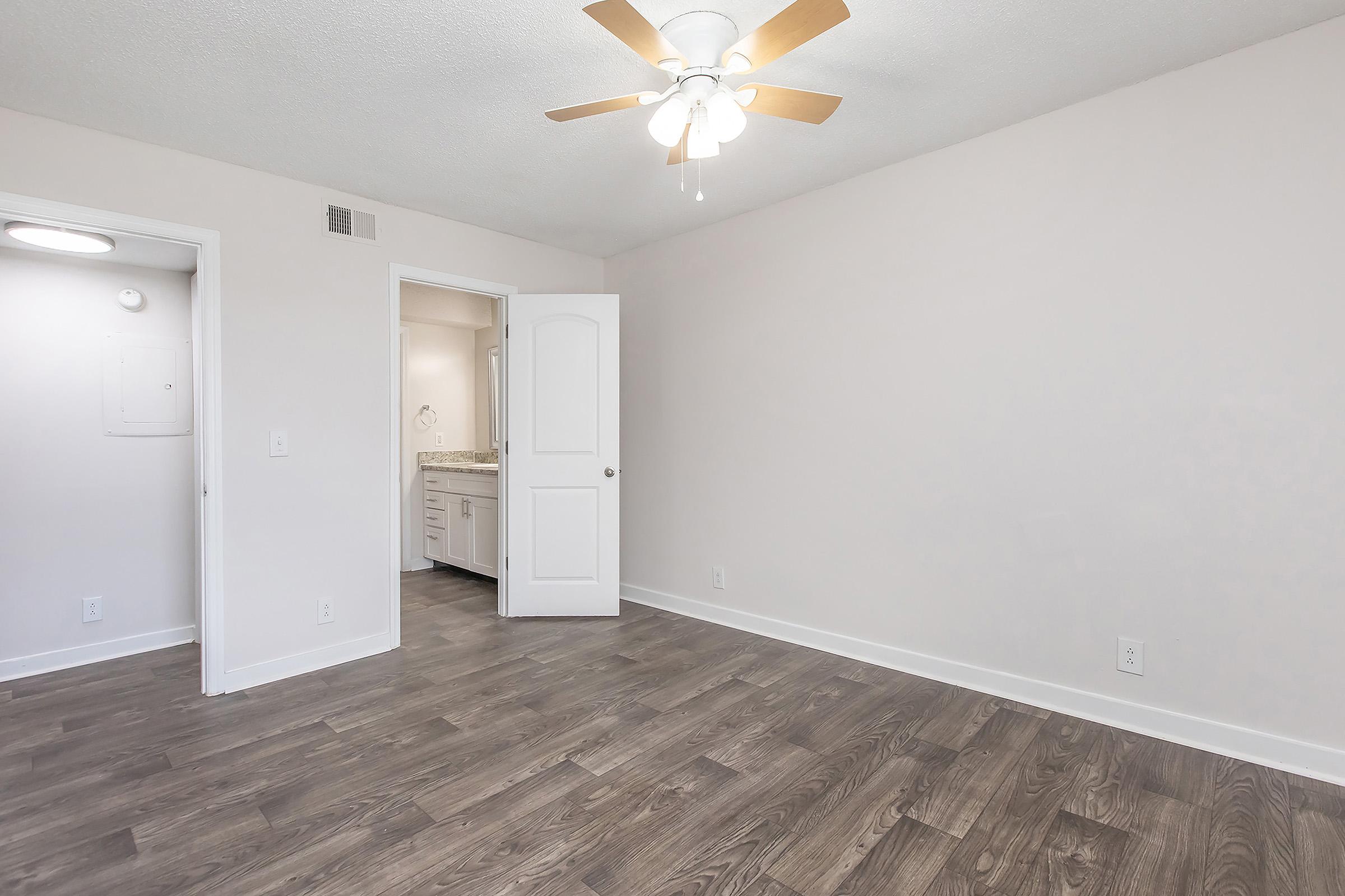 A spacious room featuring light-colored walls, a ceiling fan with wooden blades, and dark wood-like flooring. A doorway leads to a bathroom area, with natural light enhancing the open atmosphere. The room is unadorned and ready for personal decoration, creating a blank canvas for future design.