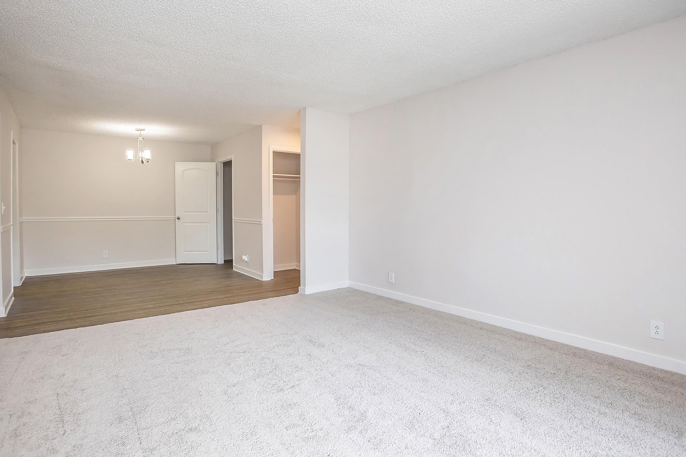 A spacious, empty room with light-colored carpet and textured white walls. To the left, a doorway leads to another room with a chandelier and a closet. The space is bright and inviting, perfect for various furniture arrangements. The clean and neutral design offers a versatile living area.
